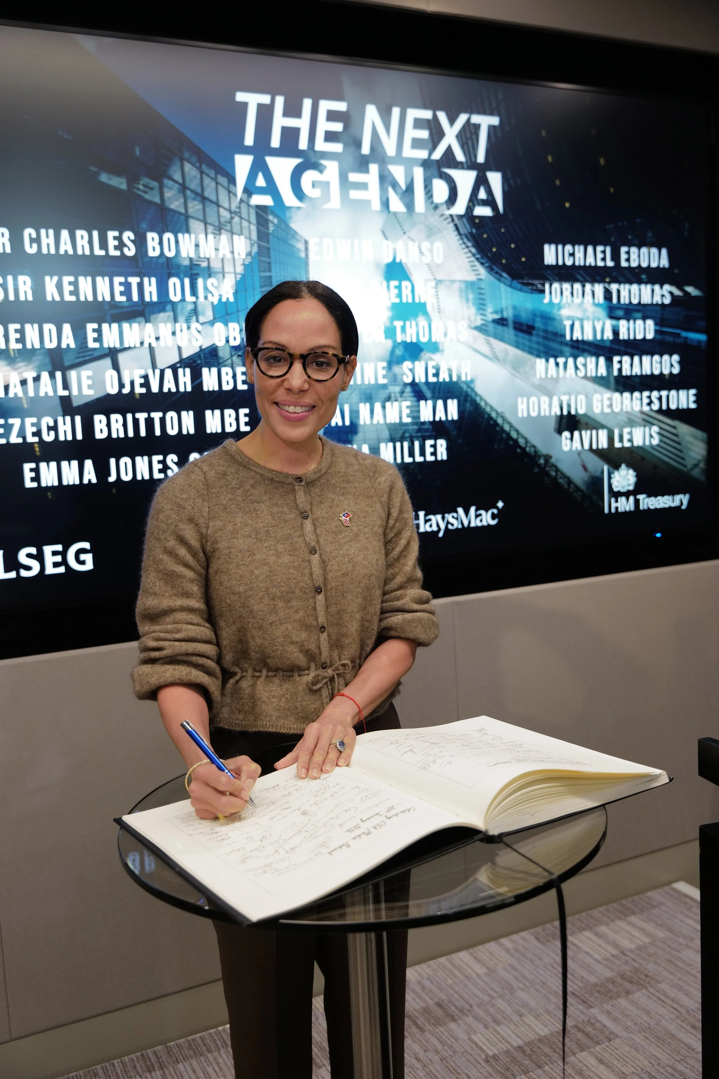 A woman wearing glasses and a brown cardigan signing a large guestbook on a round glass table with a digital display behind her reading 'THE NEXT AGENDA' and listing names.