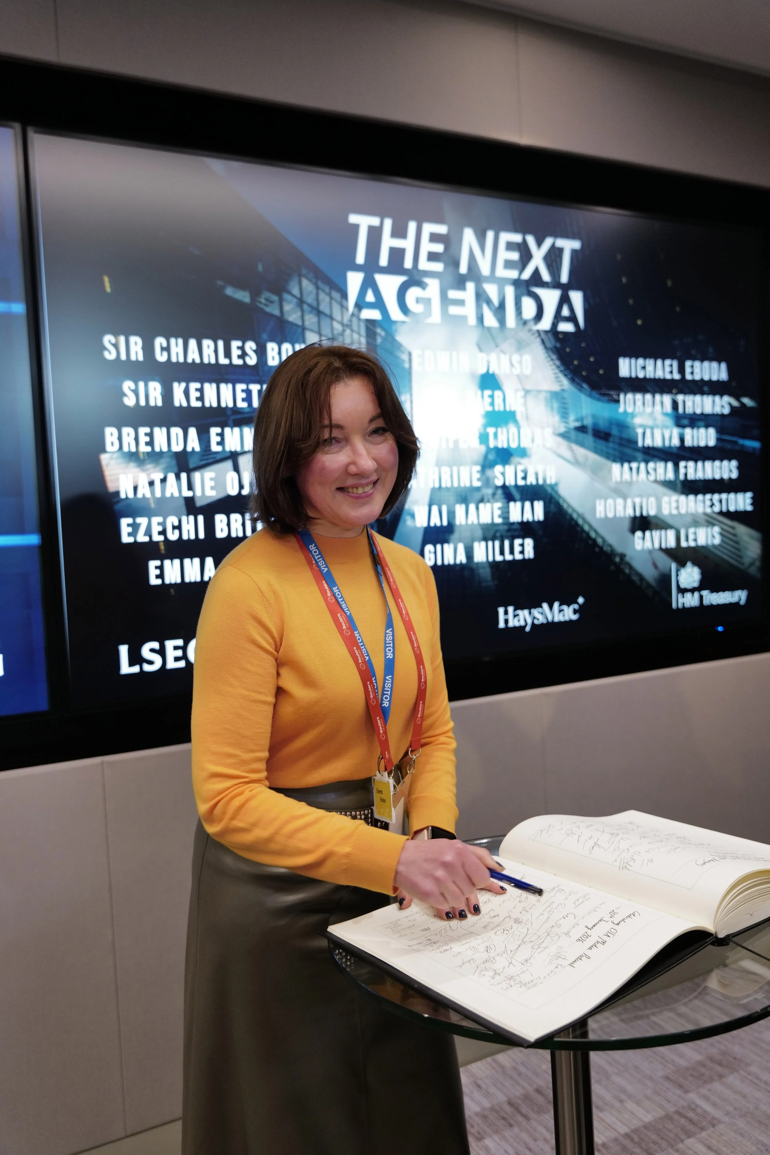 A woman in a yellow sweater signs a large guestbook on a glass table, standing in front of a screen displaying 'The Next Agenda' and multiple names.