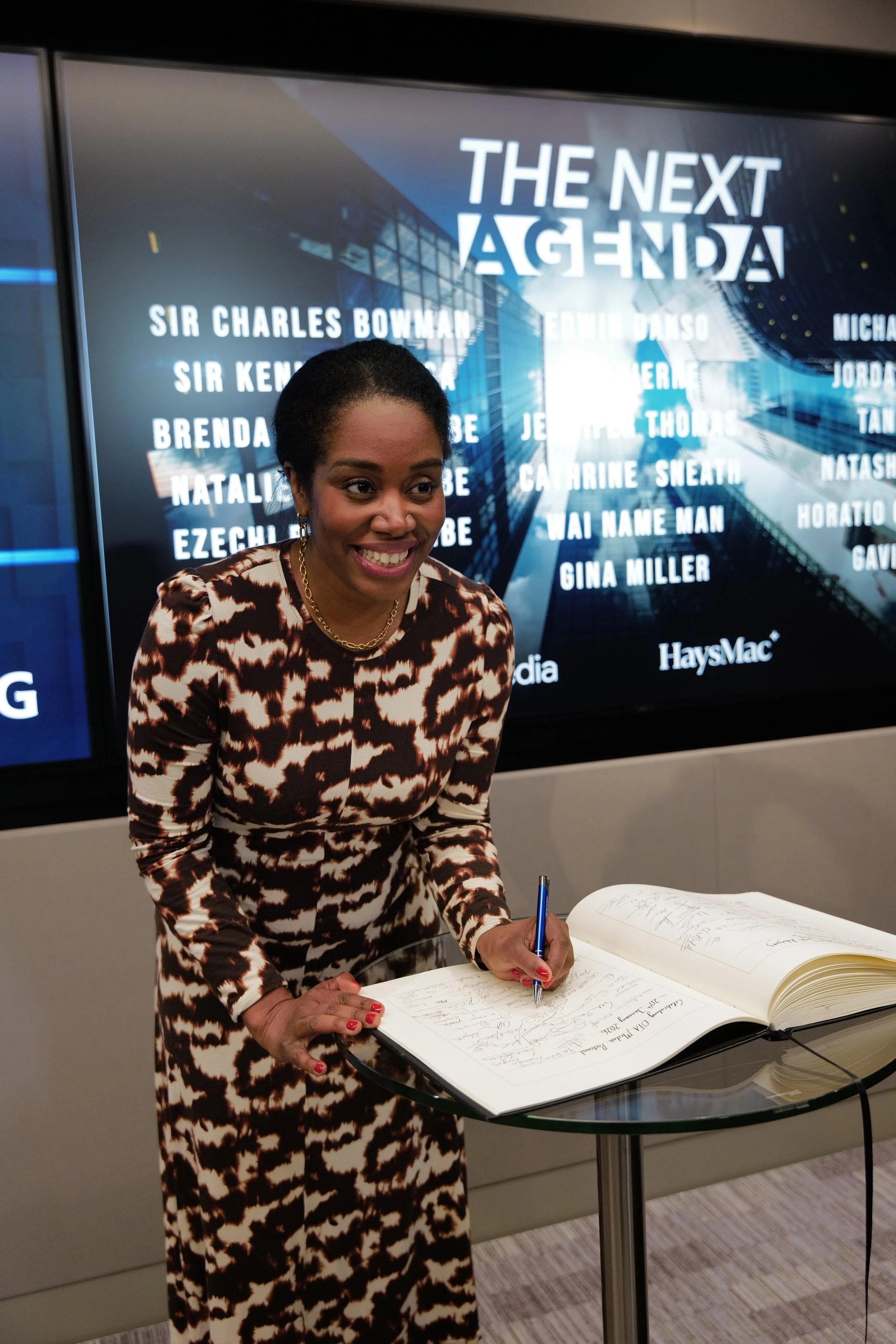 Smiling woman in patterned dress signing a large book on a glass table in front of a screen displaying the text 'THE NEXT AGENDA' and various names.