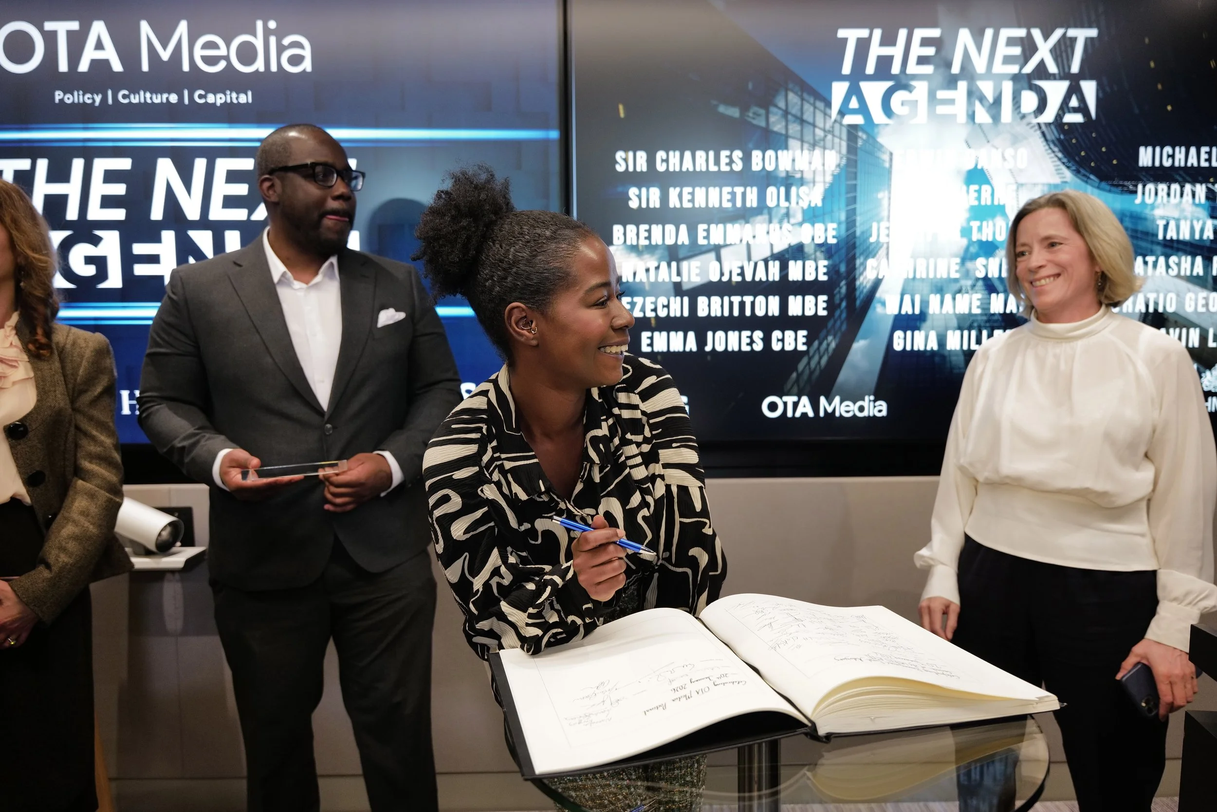 A smiling woman signs a large book at a media event while two people stand nearby with screens displaying 'The Next Agenda' and a list of names behind them.