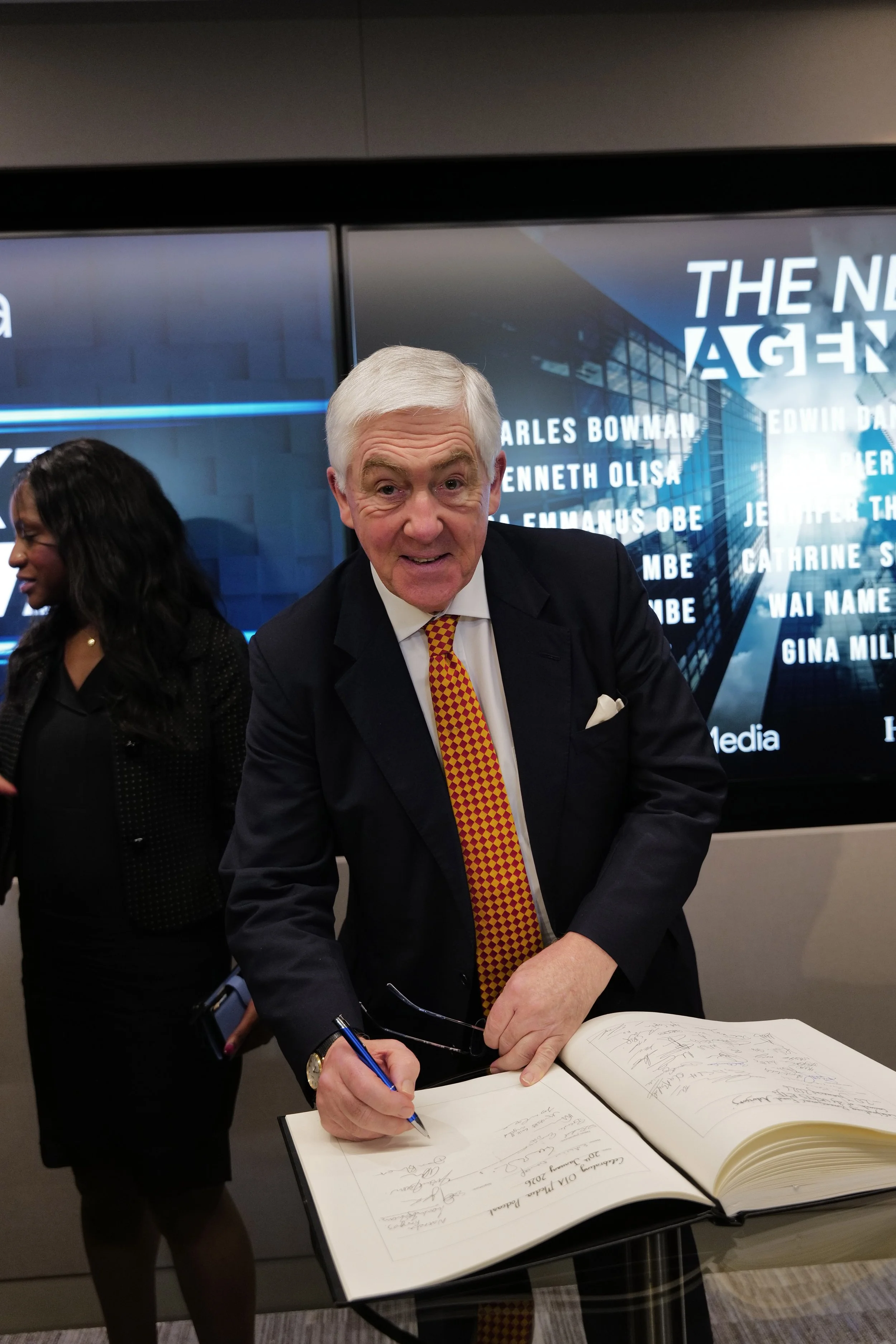 Older man in a suit with a yellow and red checkered tie signing a large book on a glass table, with a woman standing nearby and a digital screen in the background.
