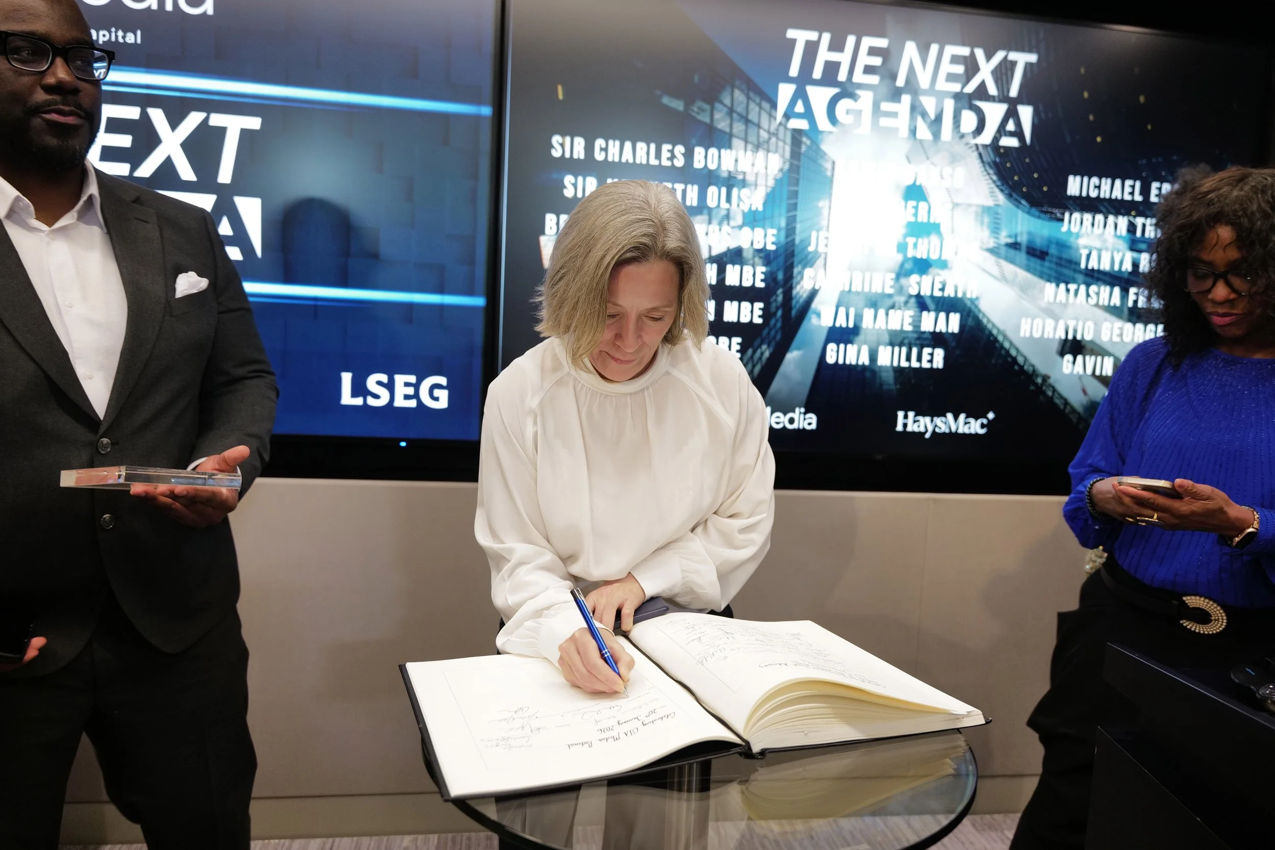 A woman in a white blouse signing a large book on a glass table, flanked by a man in a dark suit holding an award and a woman in a blue top looking at her phone, with a screen behind displaying the text 'THE NEXT AGENDA'.