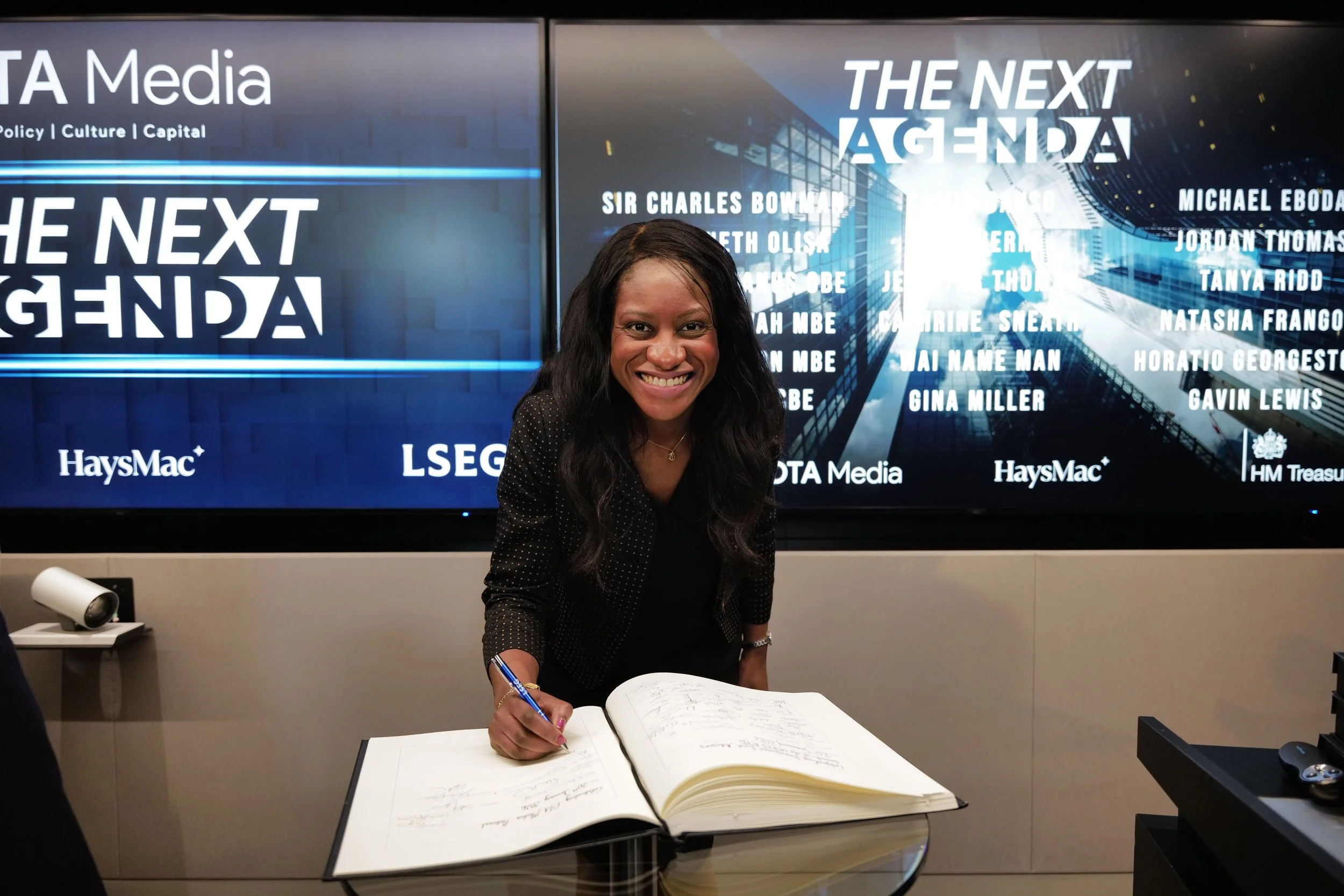 Smiling woman signing a large book at a glass table with a digital screen behind displaying 'THE NEXT AGENDA' and sponsor names.