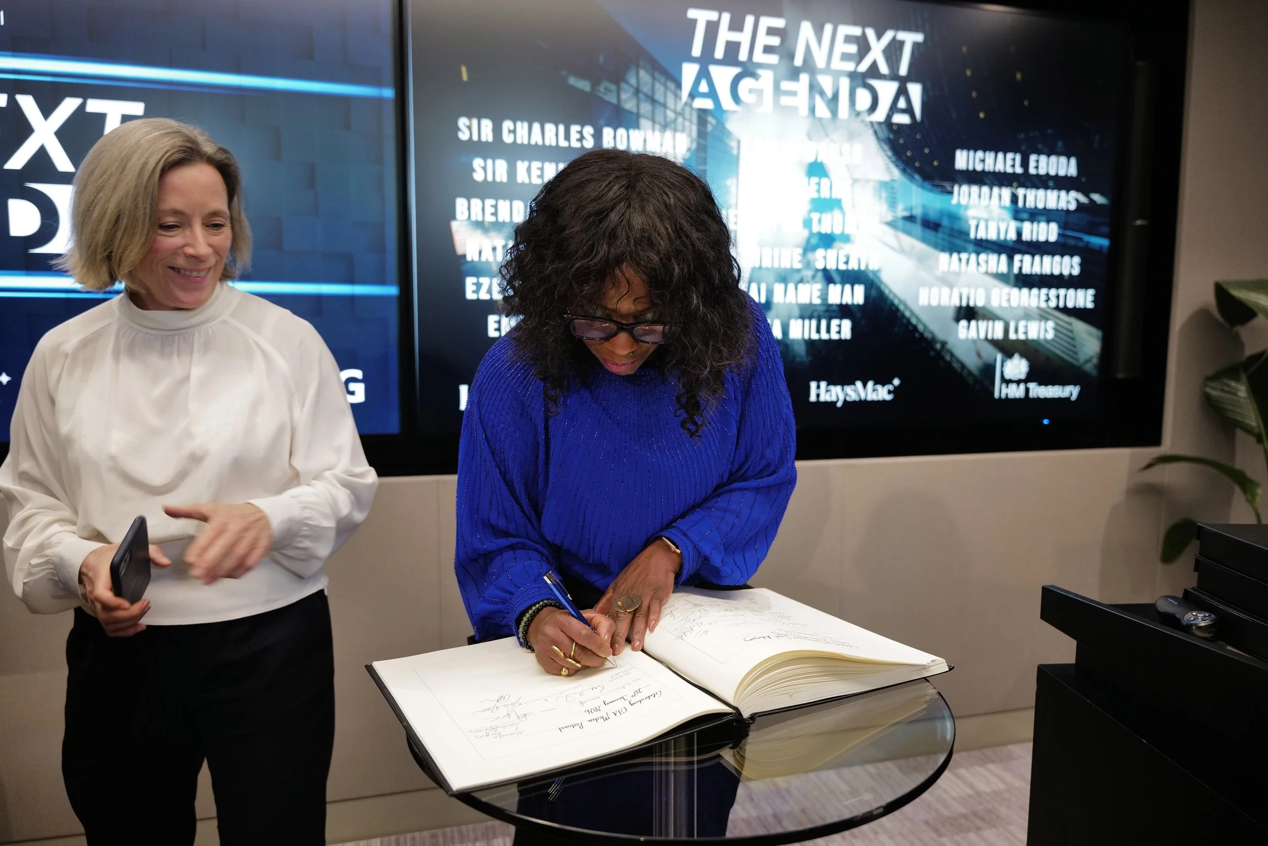 A woman in a blue sweater signs a large book on a glass table while another woman in a white blouse smiles nearby, with a screen behind them displaying the title 'The Next Agenda' and a list of names.