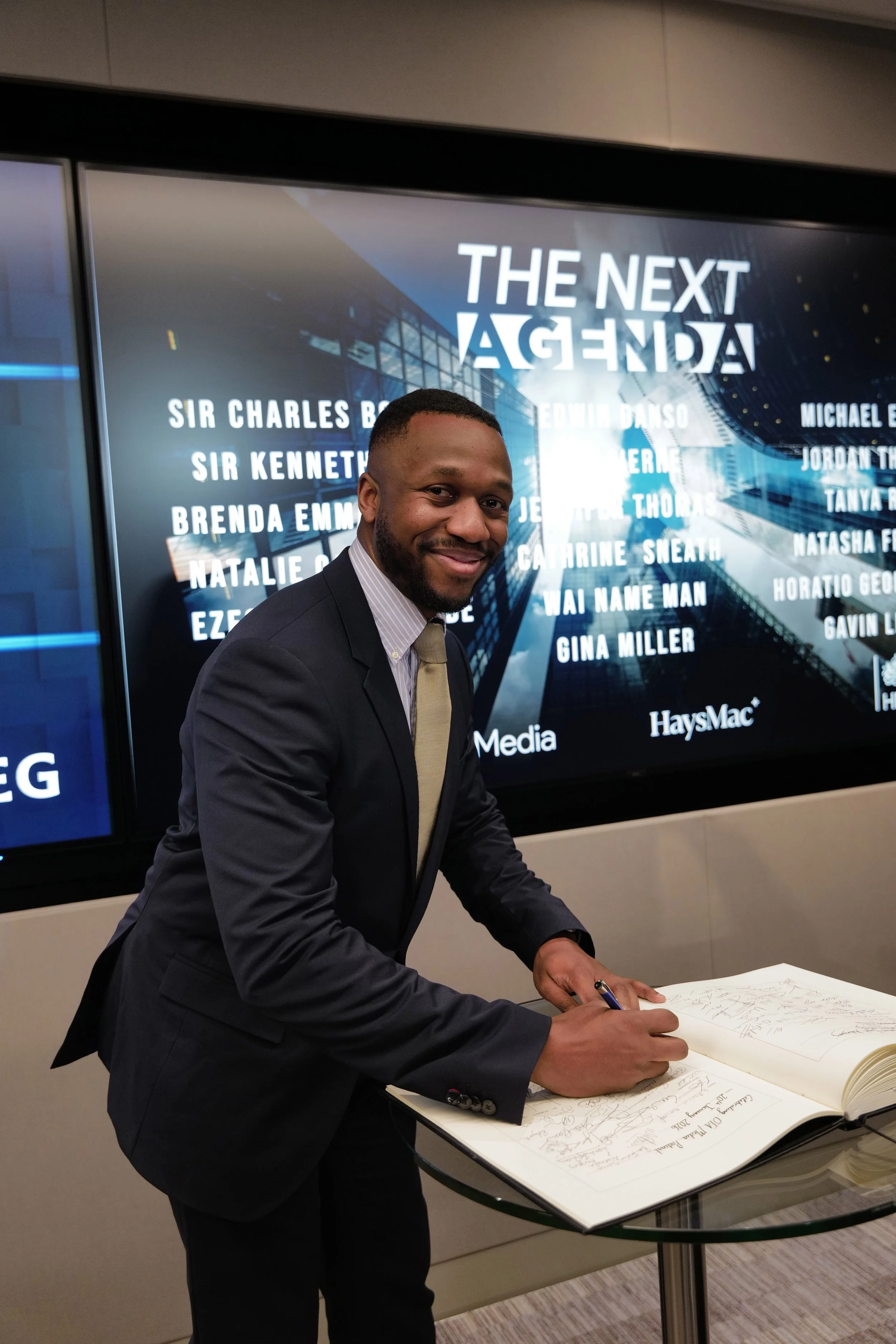 Smiling man in suit and tie signing a large book on a glass table in front of a screen displaying 'The Next Agenda' and a list of names.
