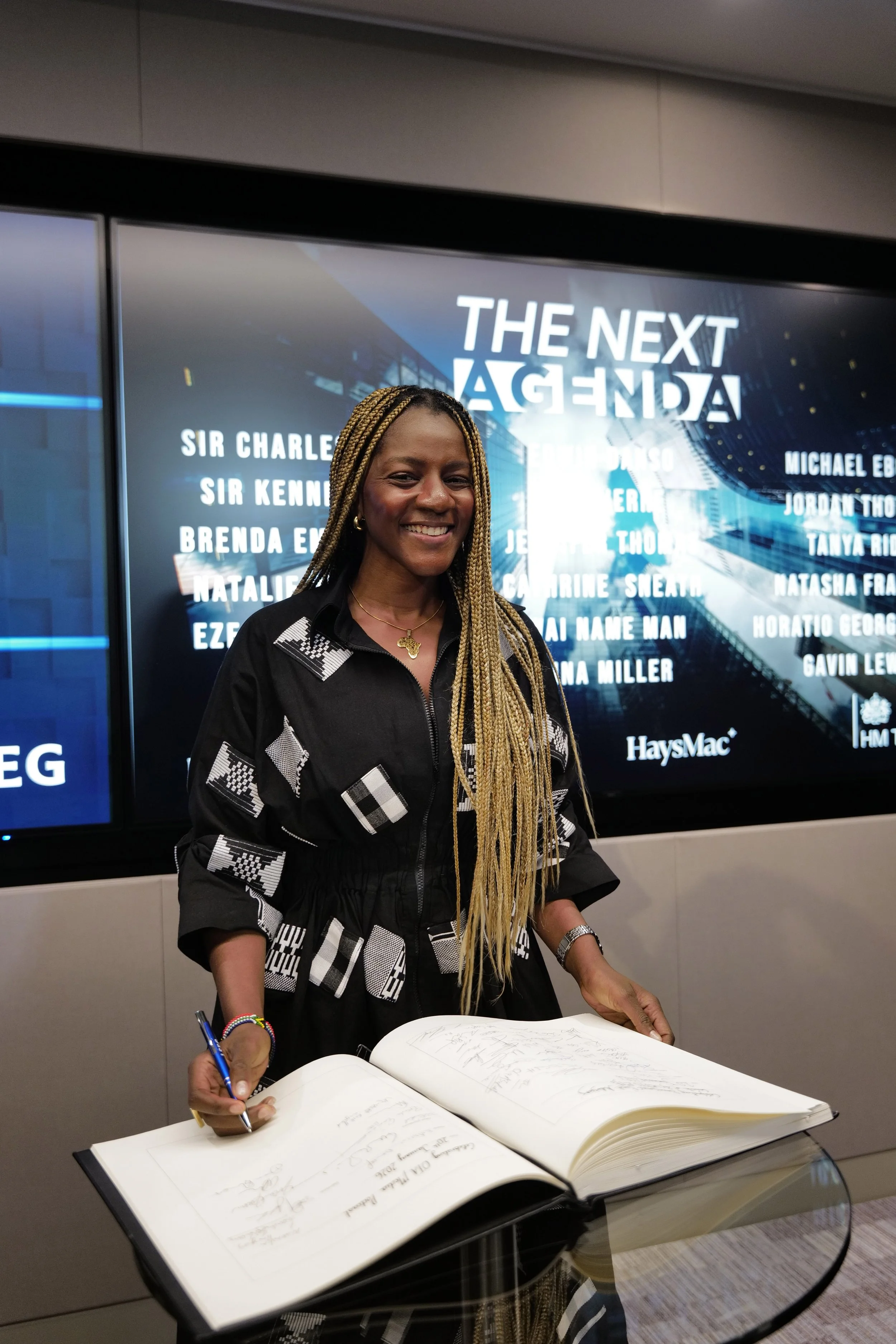 Smiling woman with long braided hair signing a large book in front of a screen displaying 'THE NEXT AGENDA' and a list of names.