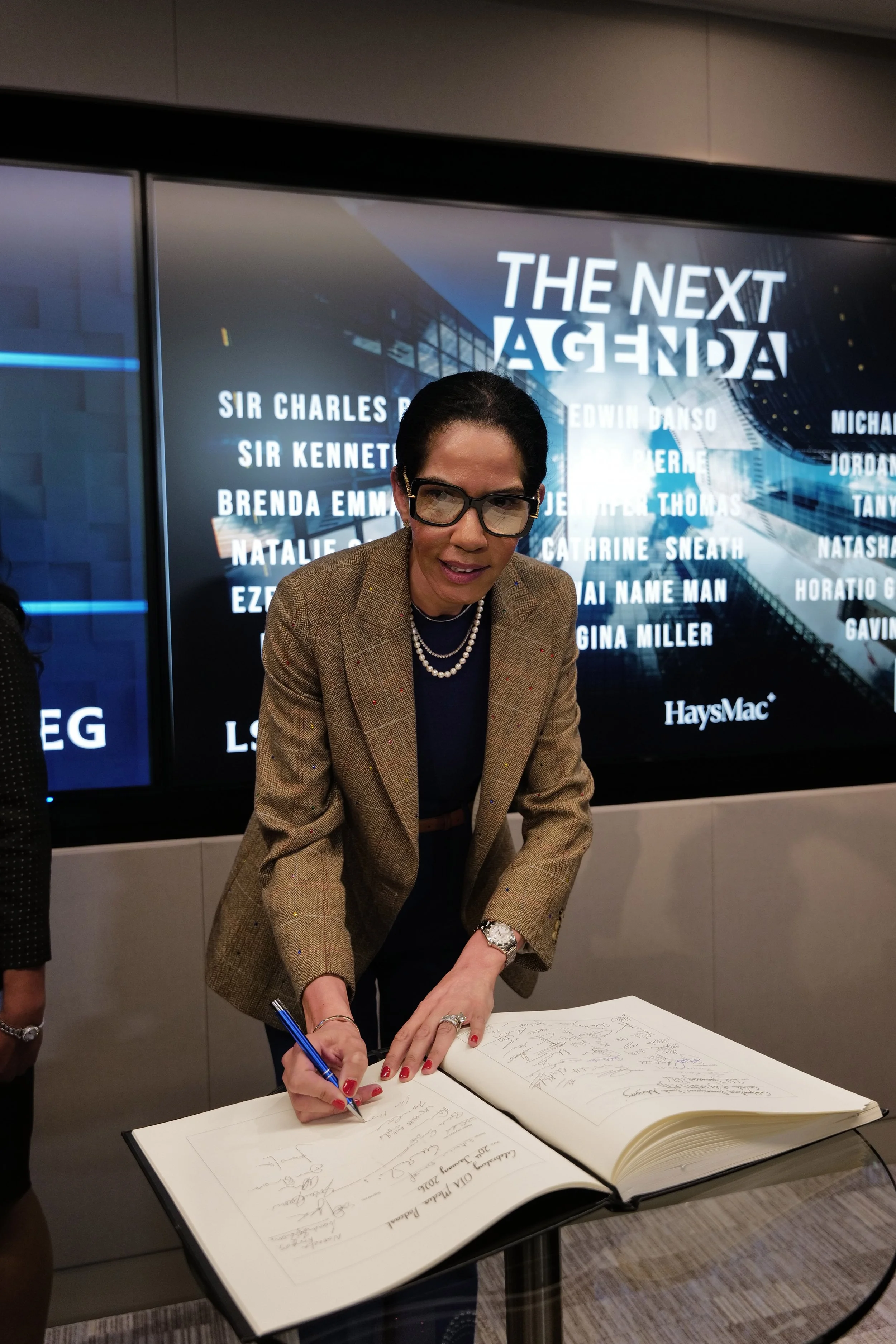 Woman wearing glasses and a brown jacket signs a large book on a glass table, with a screen behind showing the text 'THE NEXT AGENDA' and a list of names.