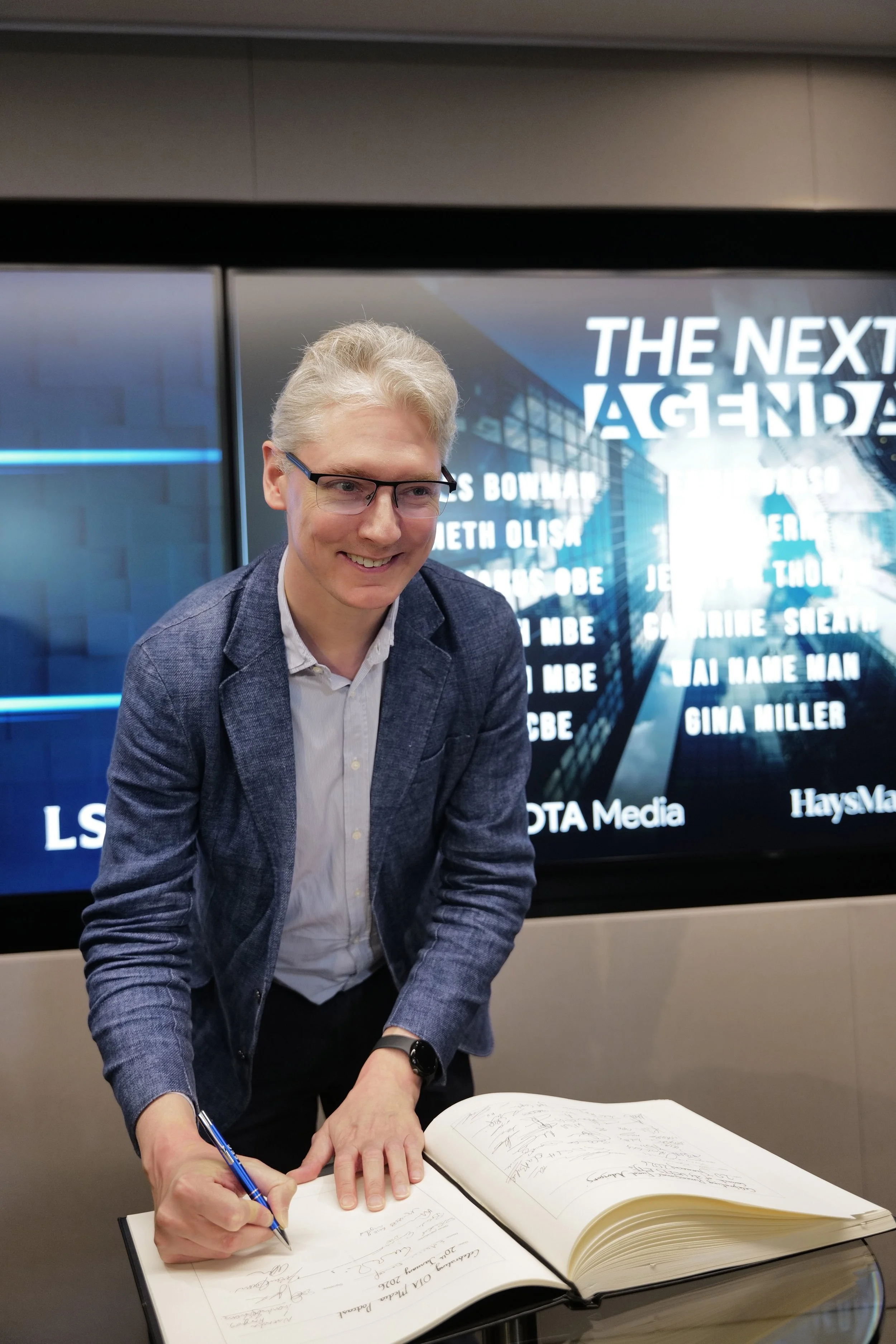 Smiling man with glasses signing a large book on a table, with a screen behind displaying 'THE NEXT AGENDA' and various names.