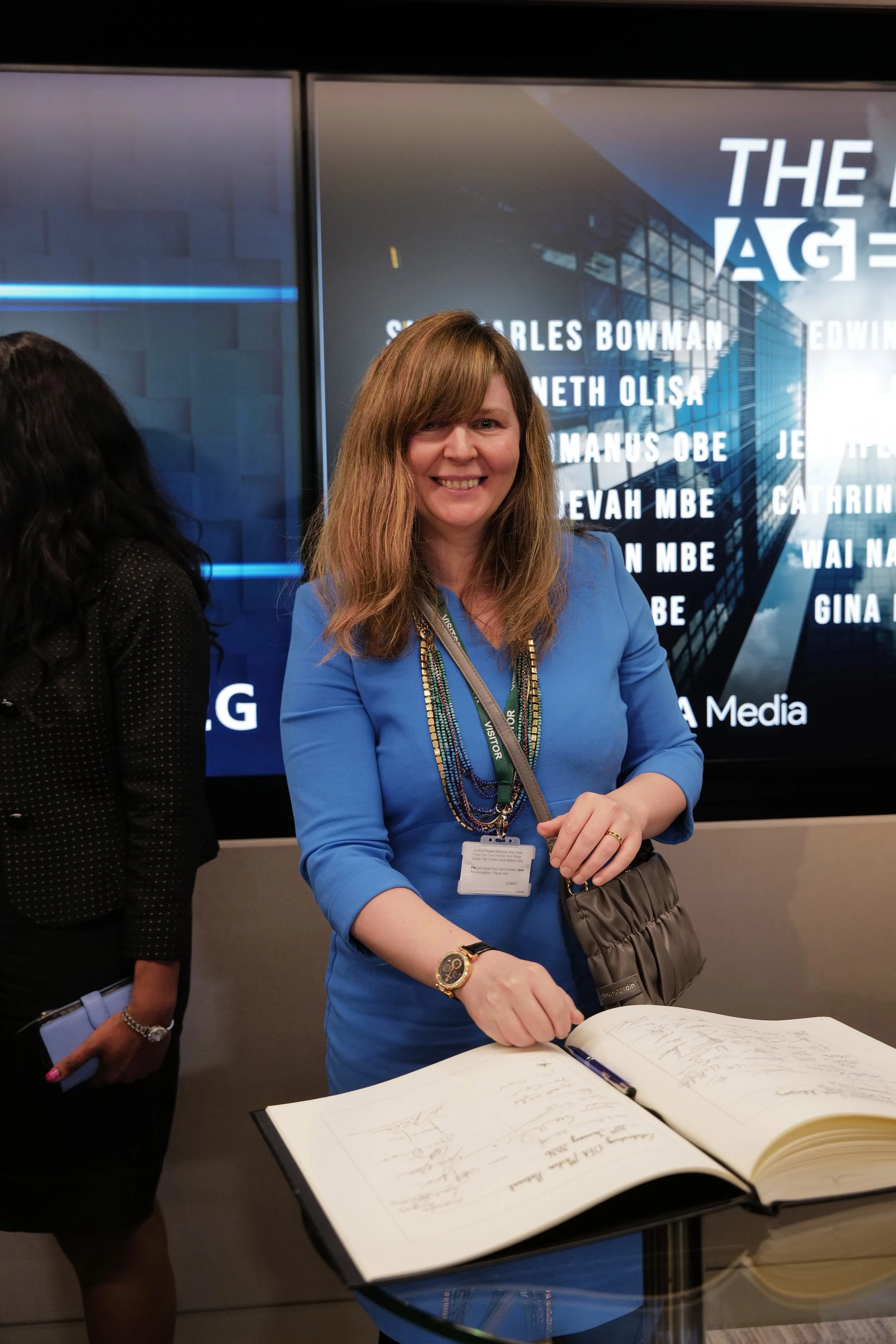 Woman in blue dress smiling while signing a large book on a glass table.