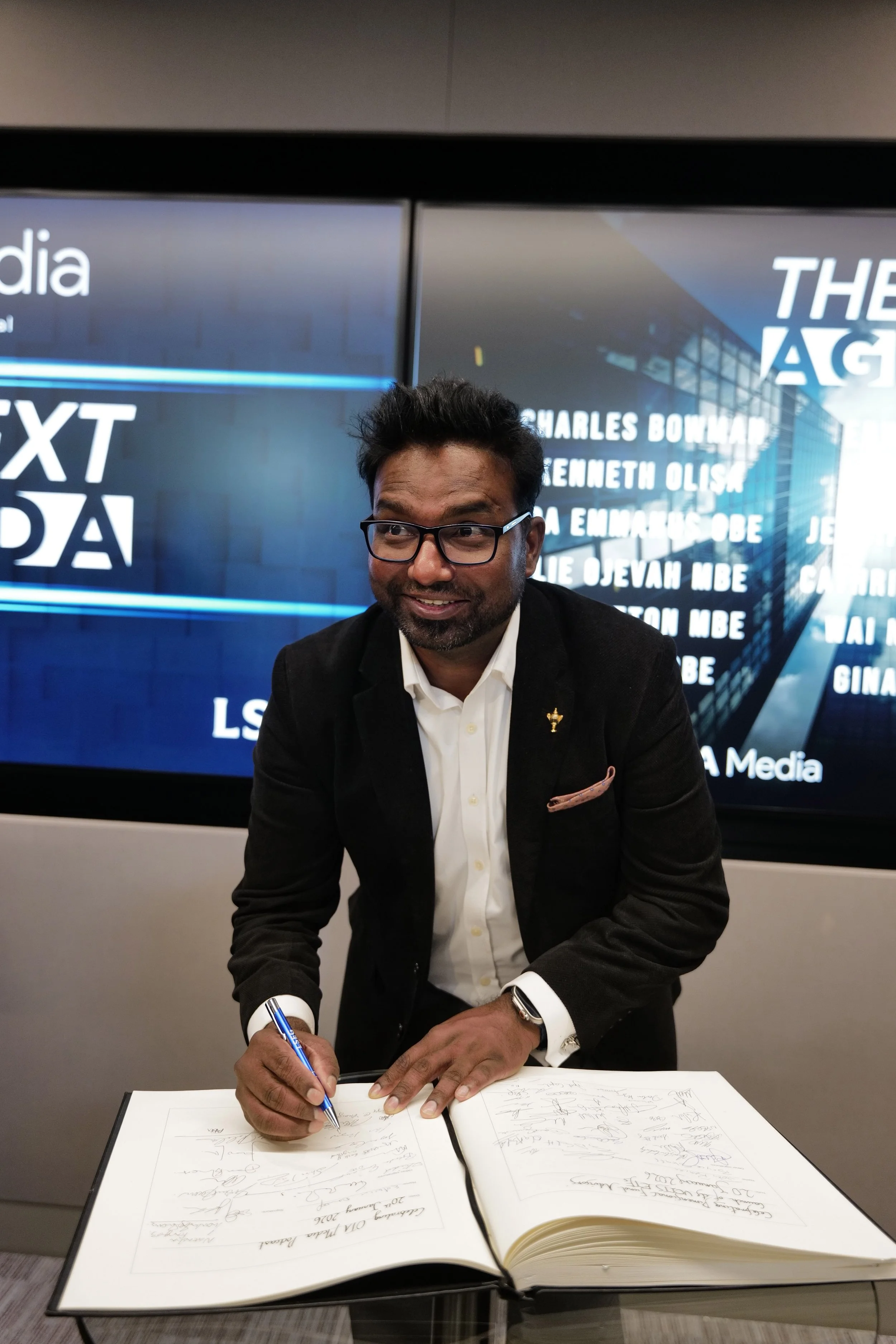 Man in glasses and black jacket signing a large book at a glass table with a digital display in the background.