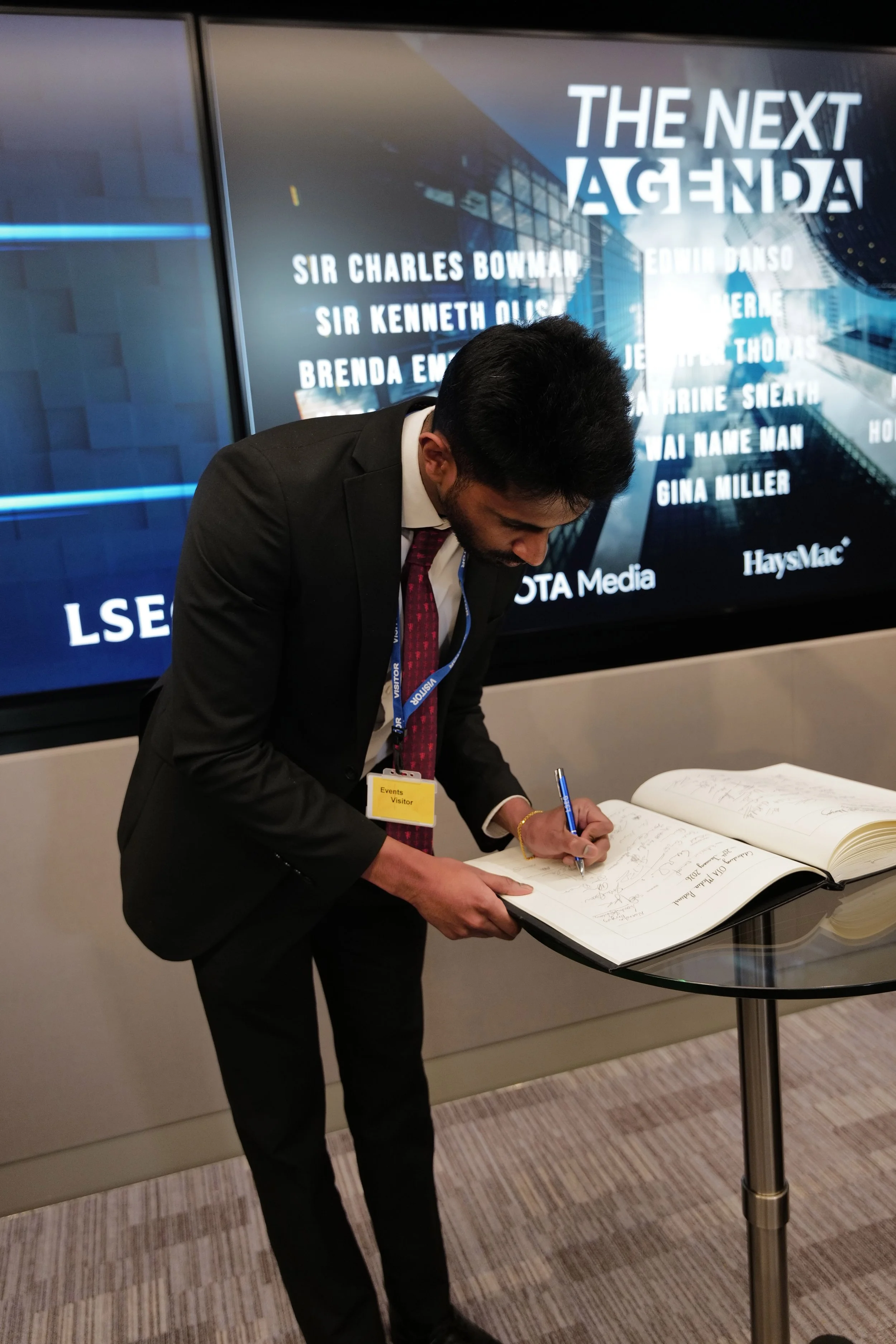 Man in a black suit and red tie signing a guest book on a round glass table in front of a screen displaying 'The Next Agenda' and a list of names.