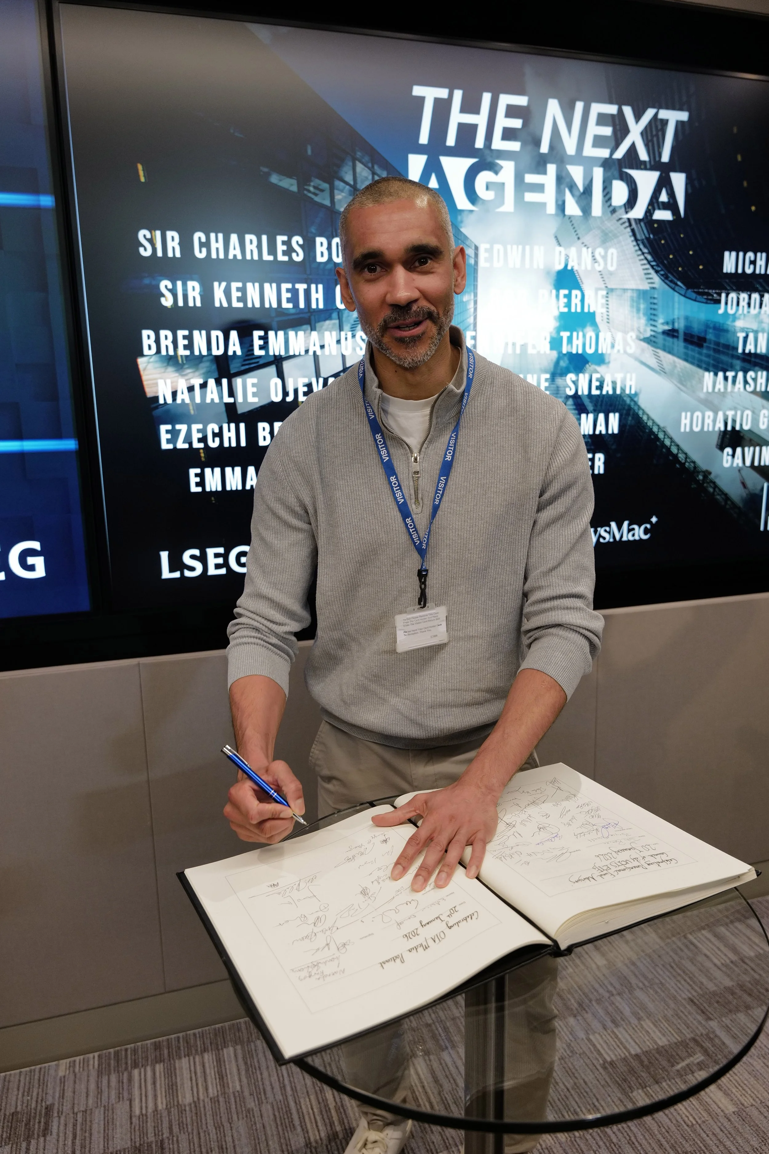 Man wearing a visitor badge signing a large book on a glass table in front of a screen displaying 'THE NEXT AGENDA' and a list of names.