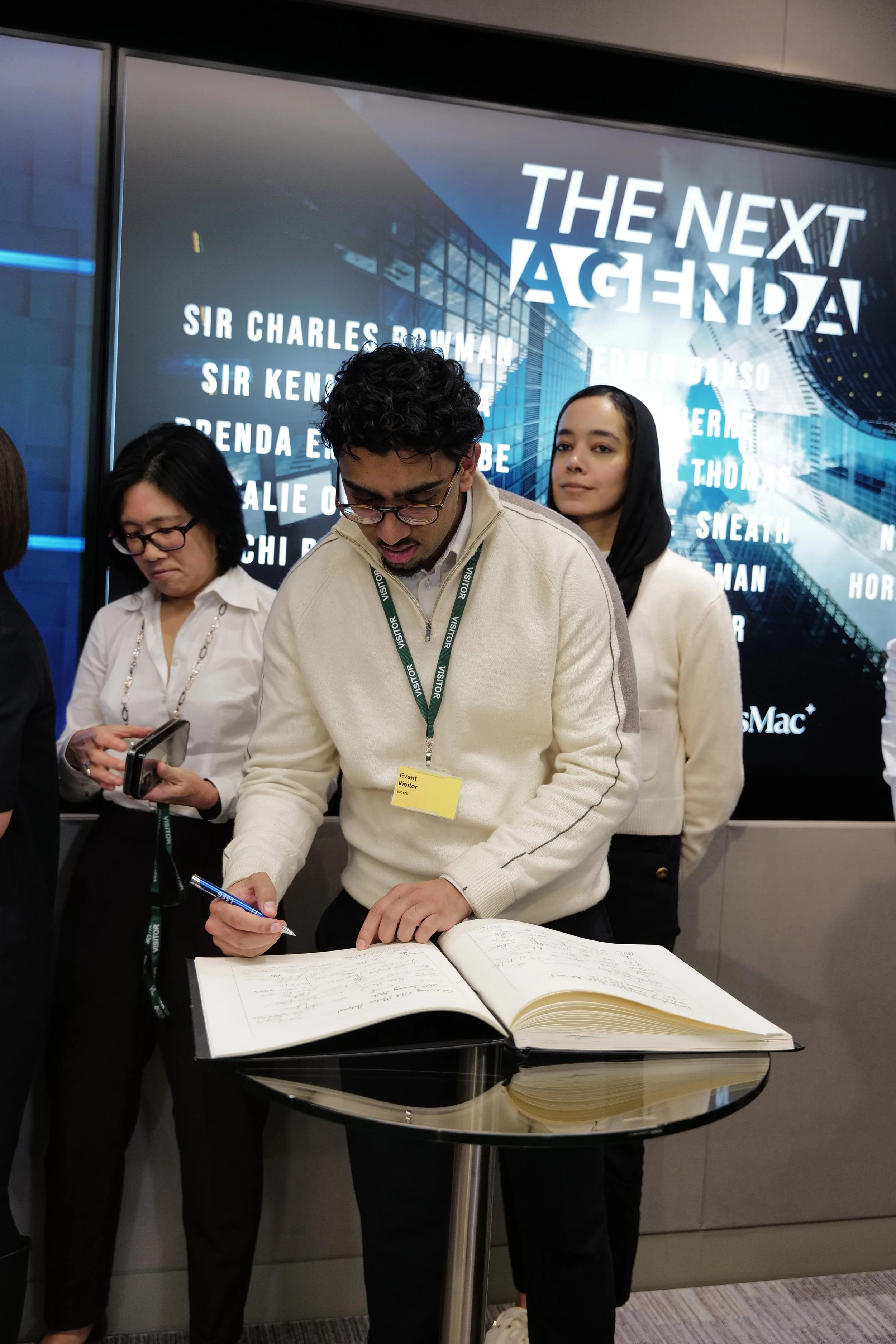 Man with glasses wearing a cream sweater signs a guestbook on a glass table while two women stand behind him.