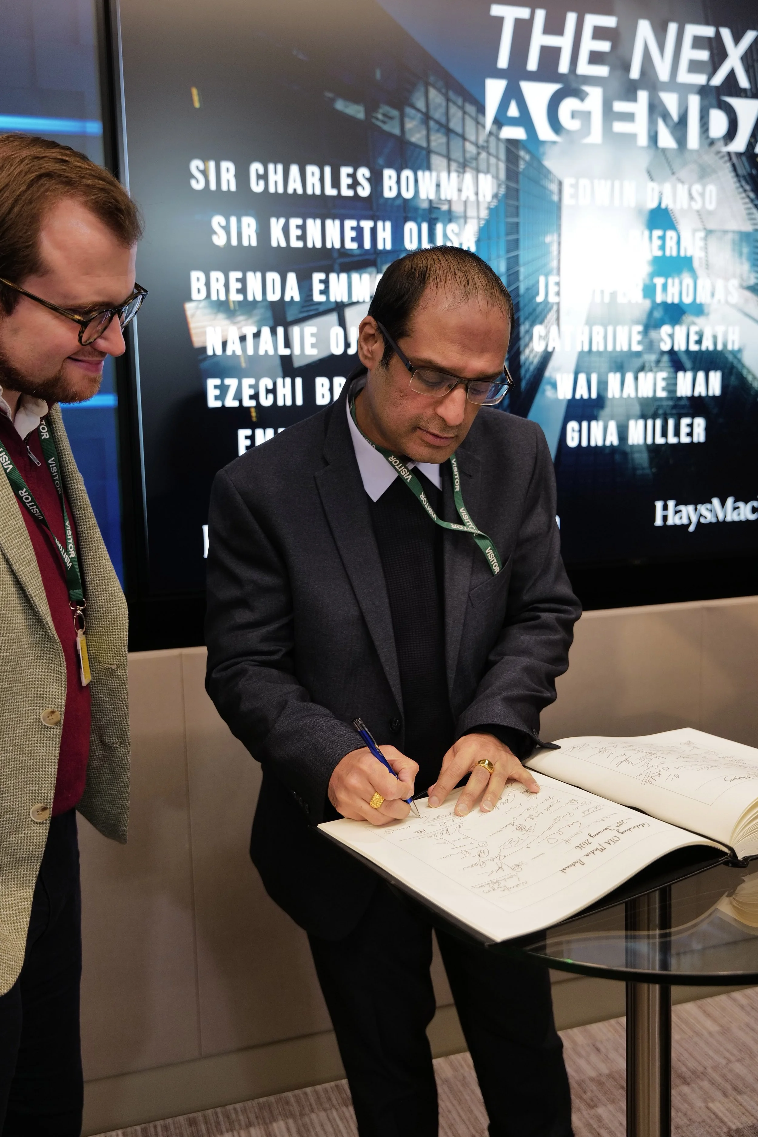 Man in glasses and dark suit signing a large guest book on a glass table while another man in a beige jacket looks on.