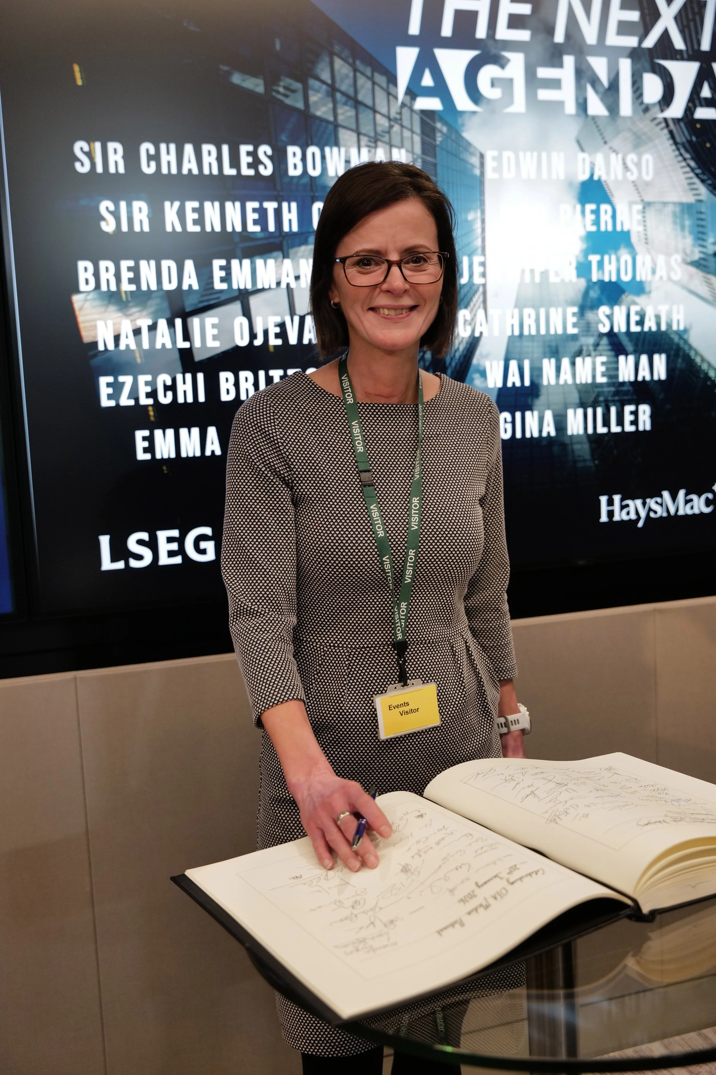 Smiling woman wearing glasses and an events visitor badge signing a large guest book at a modern venue with a digital screen showing names in the background.