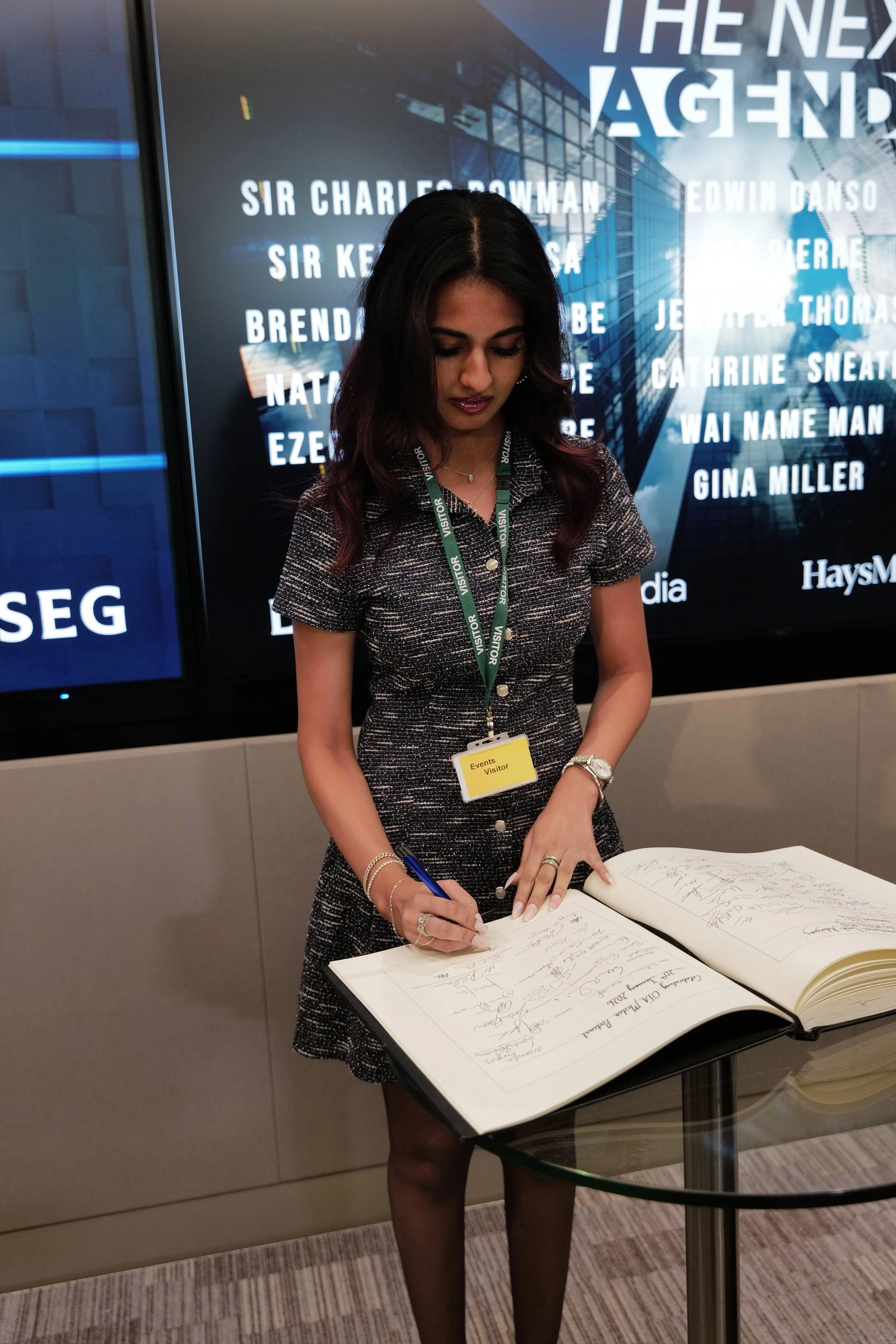 Woman wearing visitor badge signs a large guestbook on a glass table in a modern office setting.