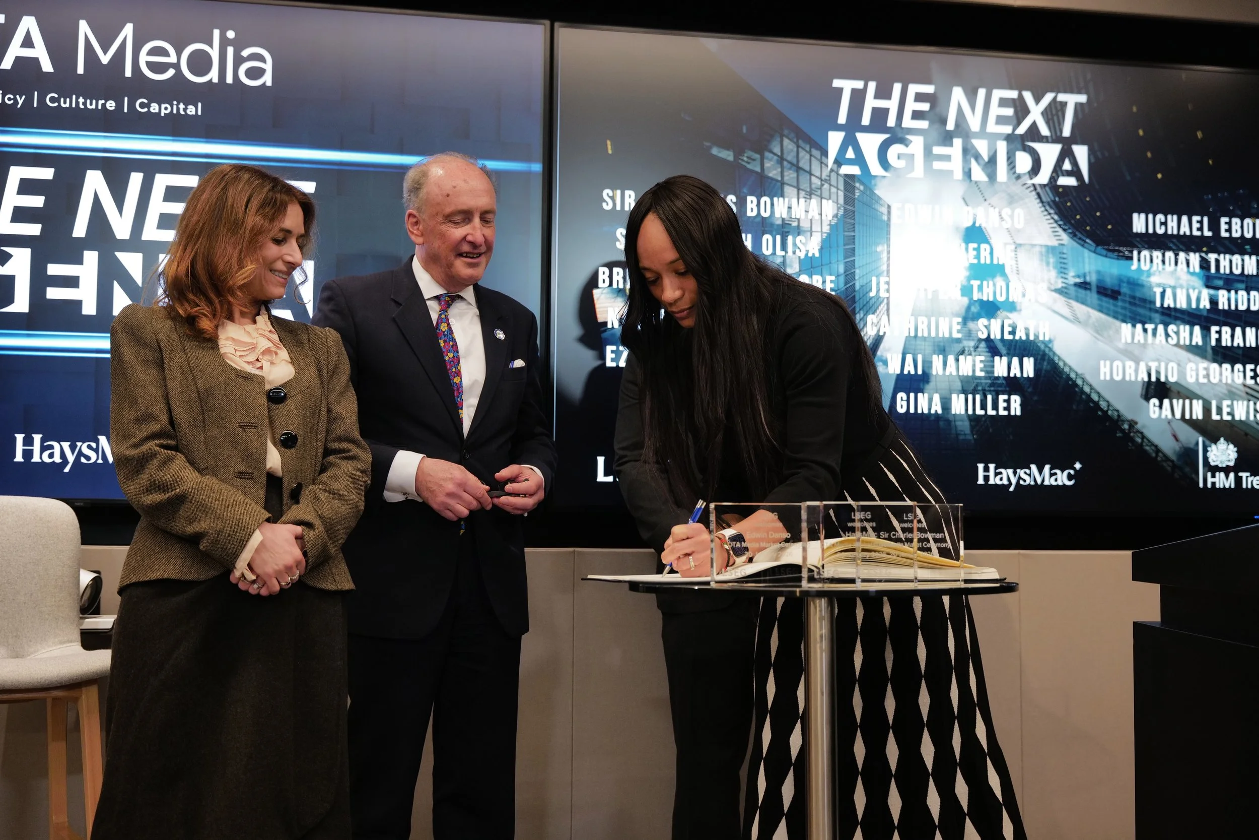 A woman in a black and white skirt signs a book on a stand while a man in a suit and a woman in a brown jacket look on, with The Next Agenda displayed on screens behind them.