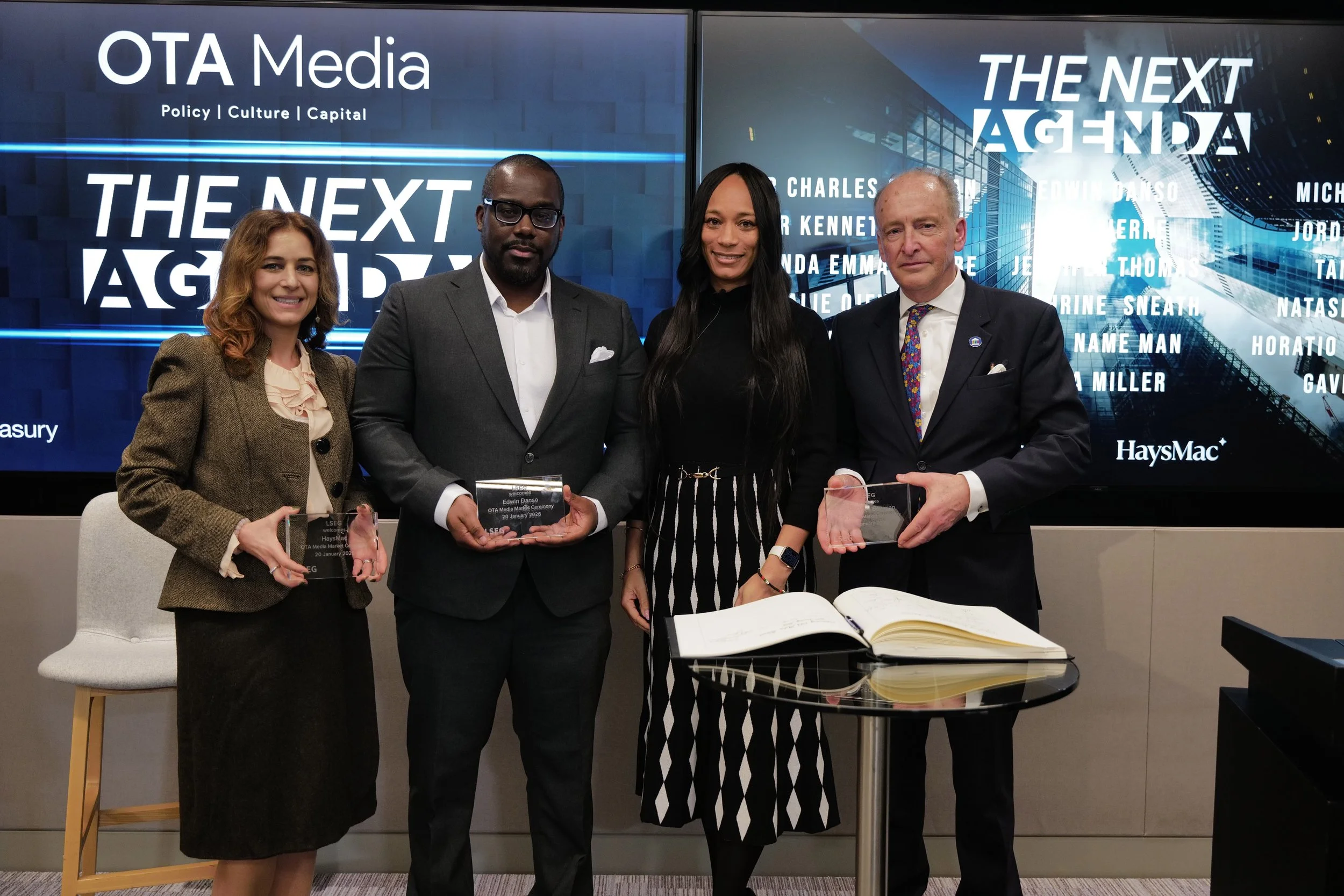 Four professionally dressed individuals standing indoors holding awards in front of screens displaying 'OTA Media' and 'The Next Agenda'.