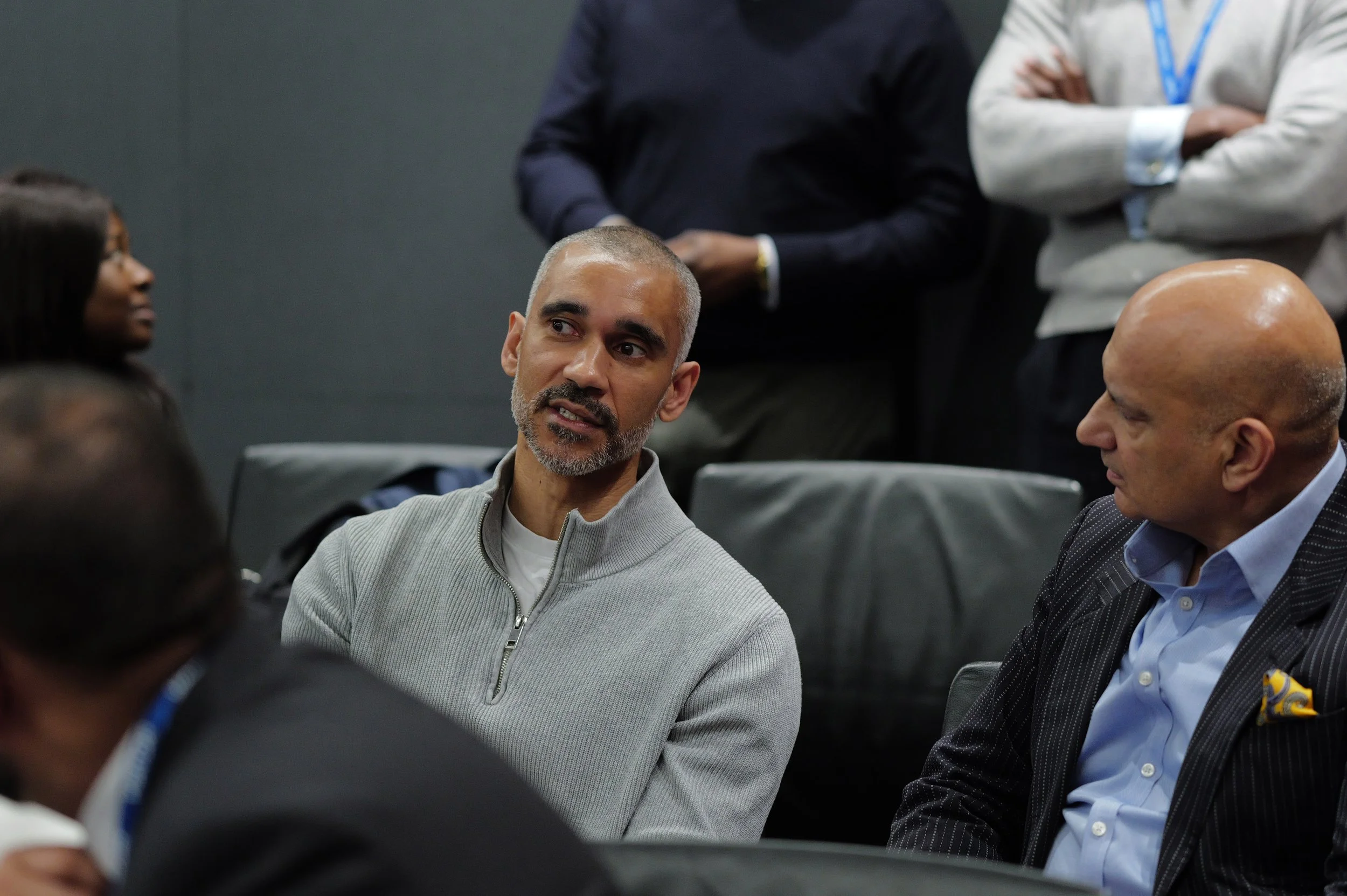 Two men seated on black leather chairs engaged in conversation, with other people blurred around them.