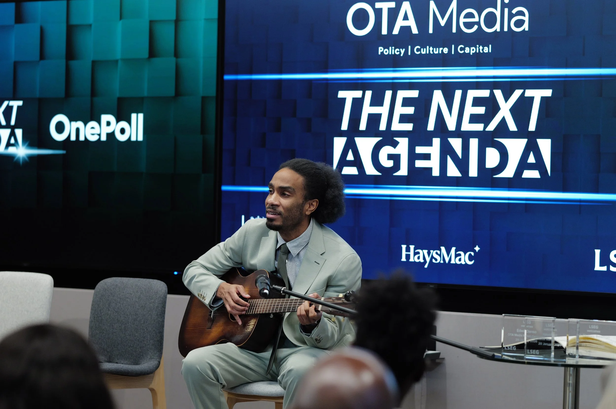 Man in a light green suit playing an acoustic guitar and singing in front of a screen displaying 'THE NEXT AGENDA' and various sponsor logos during an event.