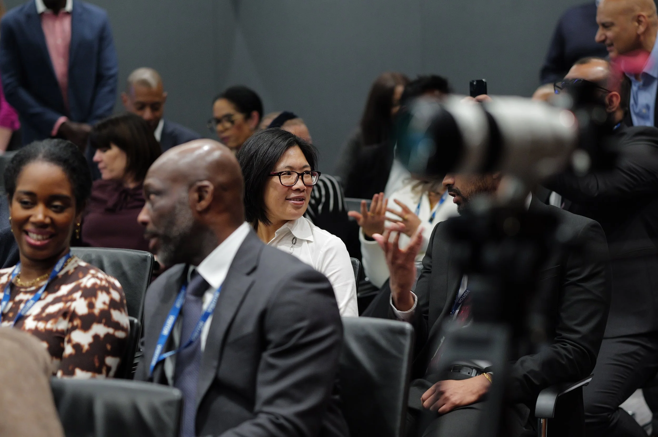 A diverse group of people in business attire sitting and engaging in conversation at a conference or event, with a camera lens prominently in the foreground.