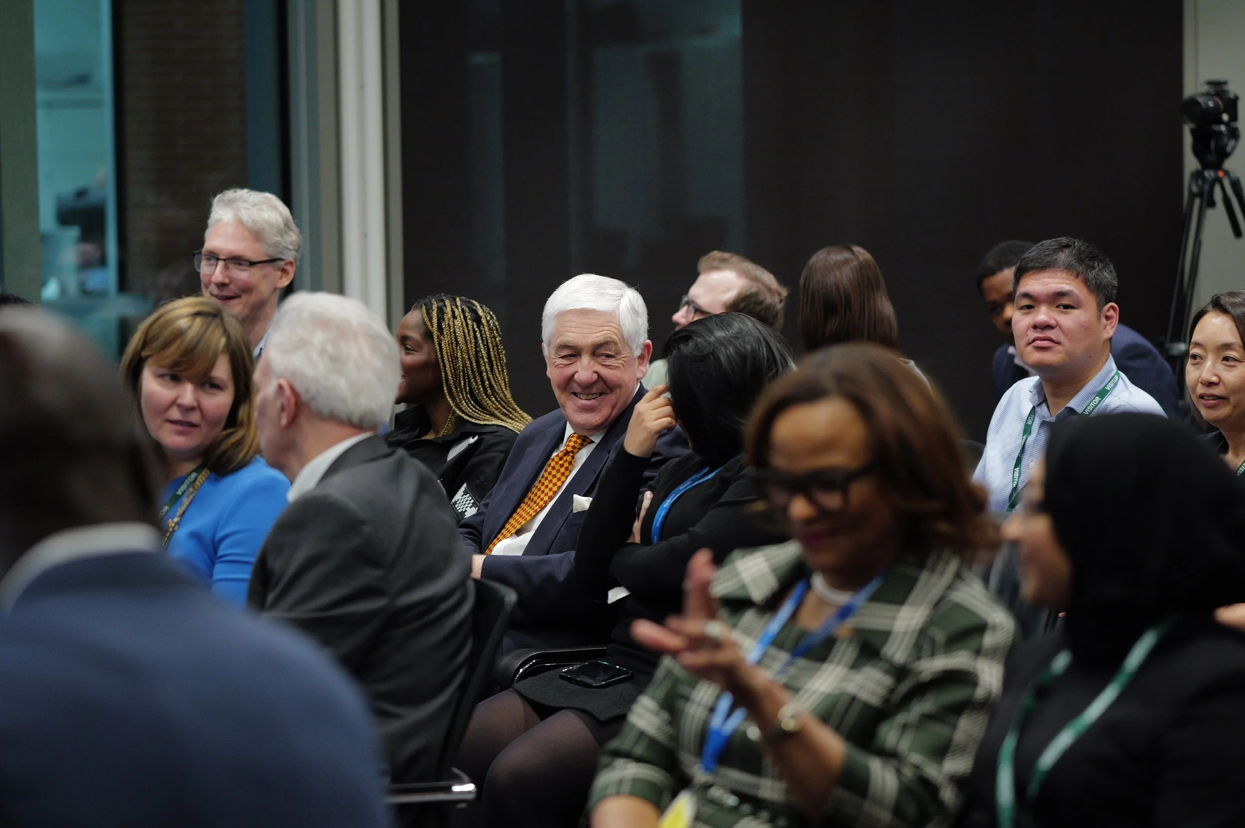 A diverse group of adults seated in rows at an indoor event, engaged in conversation and smiling.