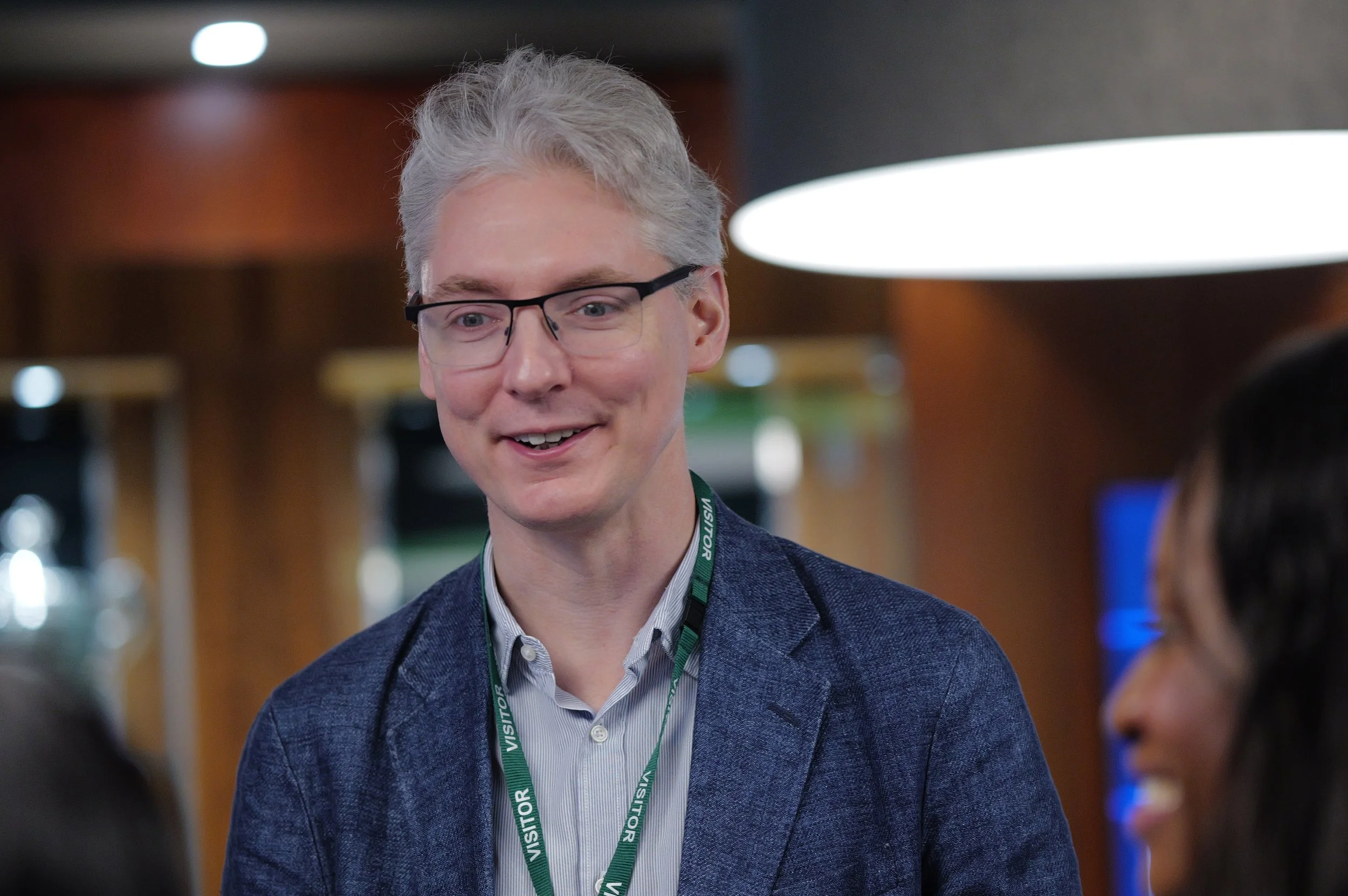 Middle-aged man with gray hair and glasses wearing a blue blazer and visitor badge, smiling indoors.