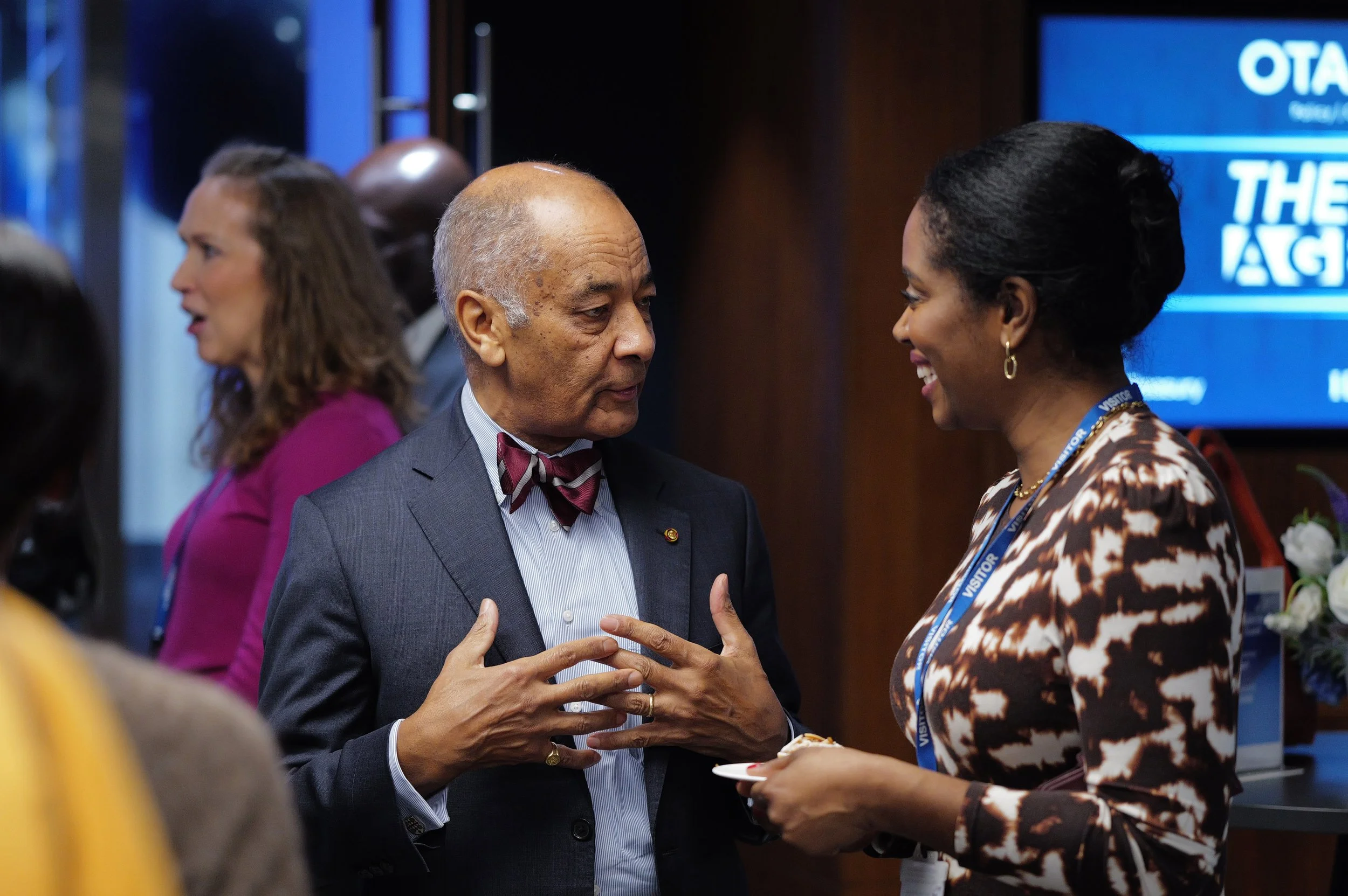 An older man in a suit and bow tie talking to a woman with a visitor badge in a patterned dress, both engaged in conversation.