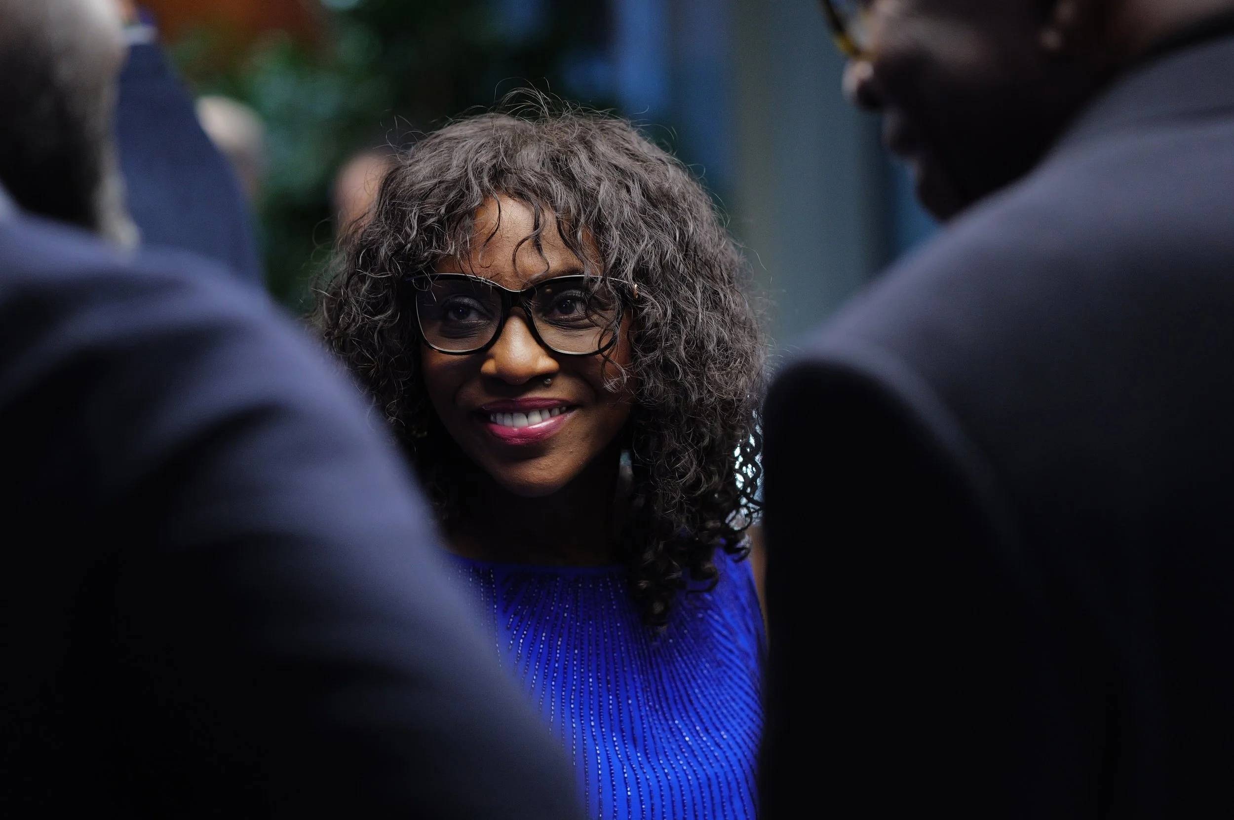 Smiling woman with curly hair and glasses wearing a blue top, engaged in conversation at a social gathering.