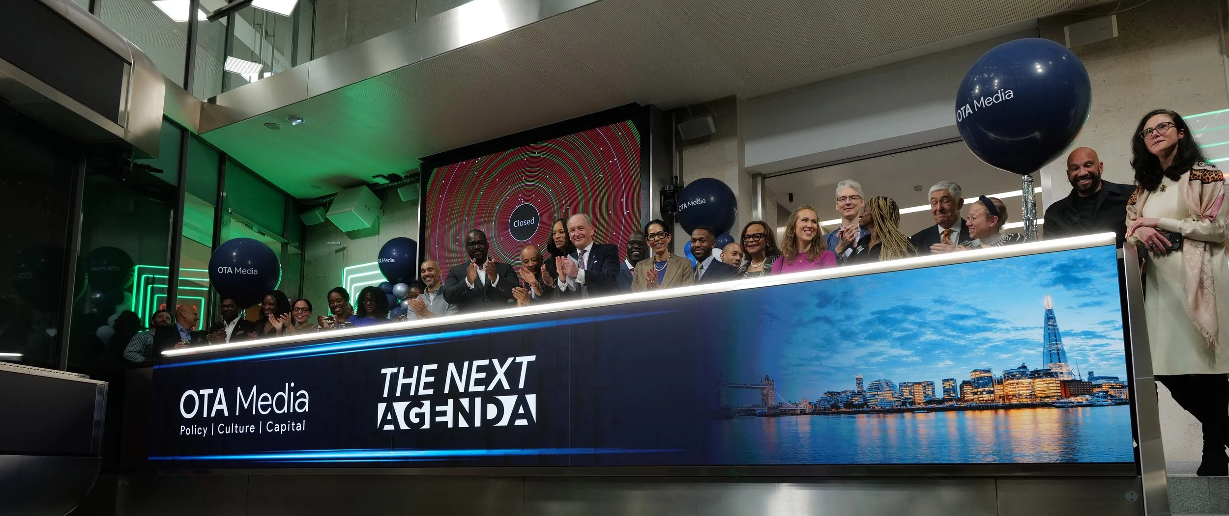 Group of people standing behind a lit counter displaying 'OTA Media Policy | Culture | Capital' and 'The Next Agenda' with a city skyline image on the right.