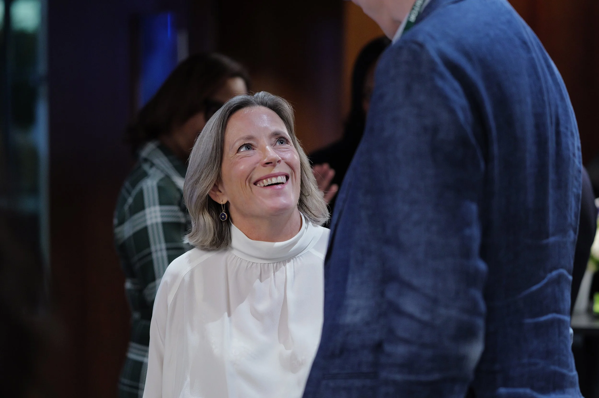 Smiling woman in white blouse looking up at a person wearing a blue jacket in a dimly lit indoor setting.