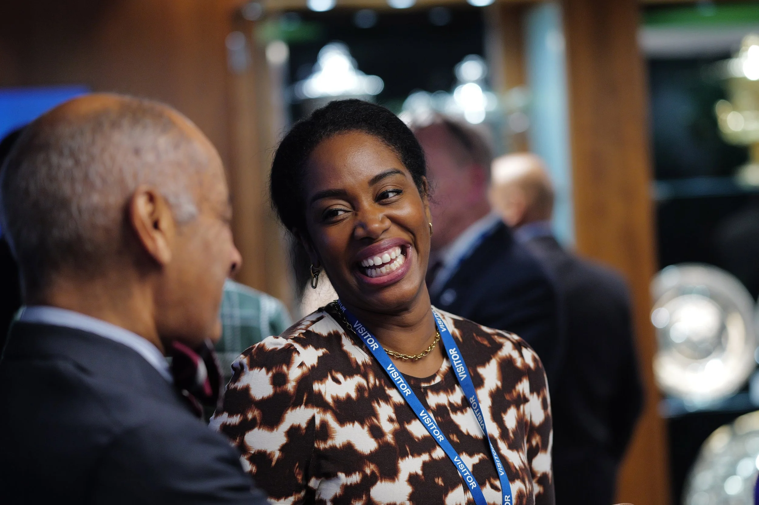Smiling woman wearing a visitor badge and patterned dress engaging in conversation with a man in a suit.