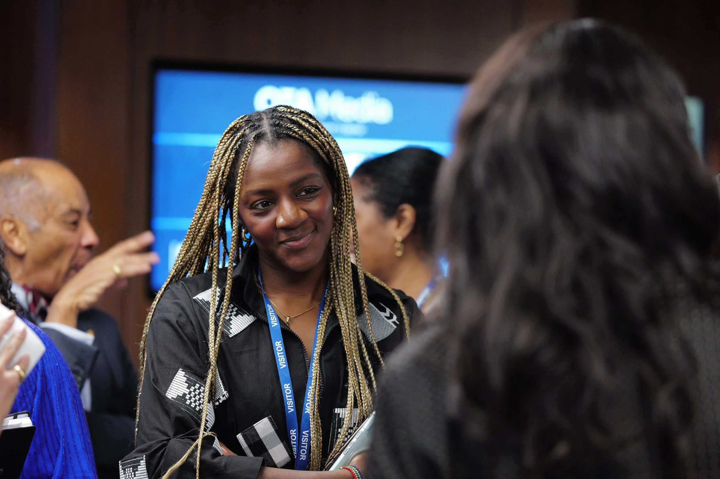 Woman with long braided hair wearing a visitor badge engaging in conversation at an indoor event.