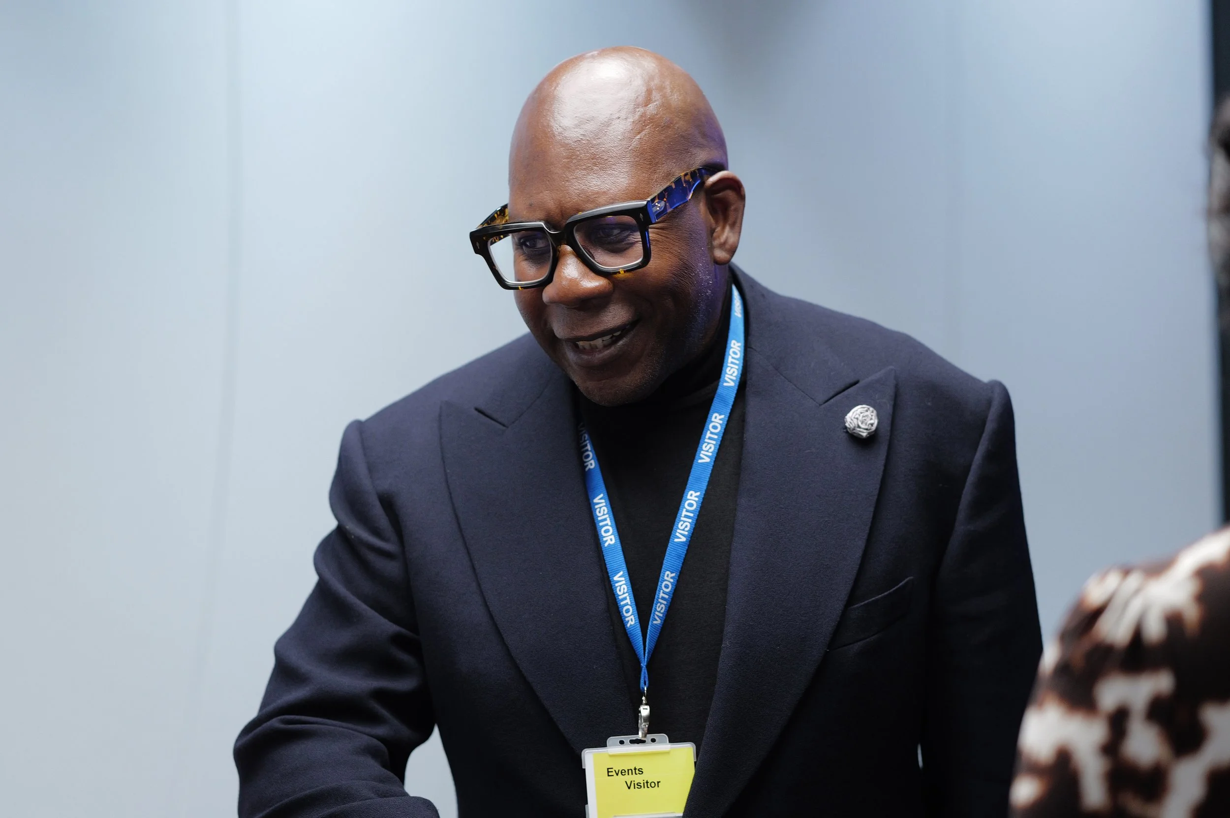 Smiling man wearing dark glasses and a black blazer with a visitor badge on a blue lanyard.