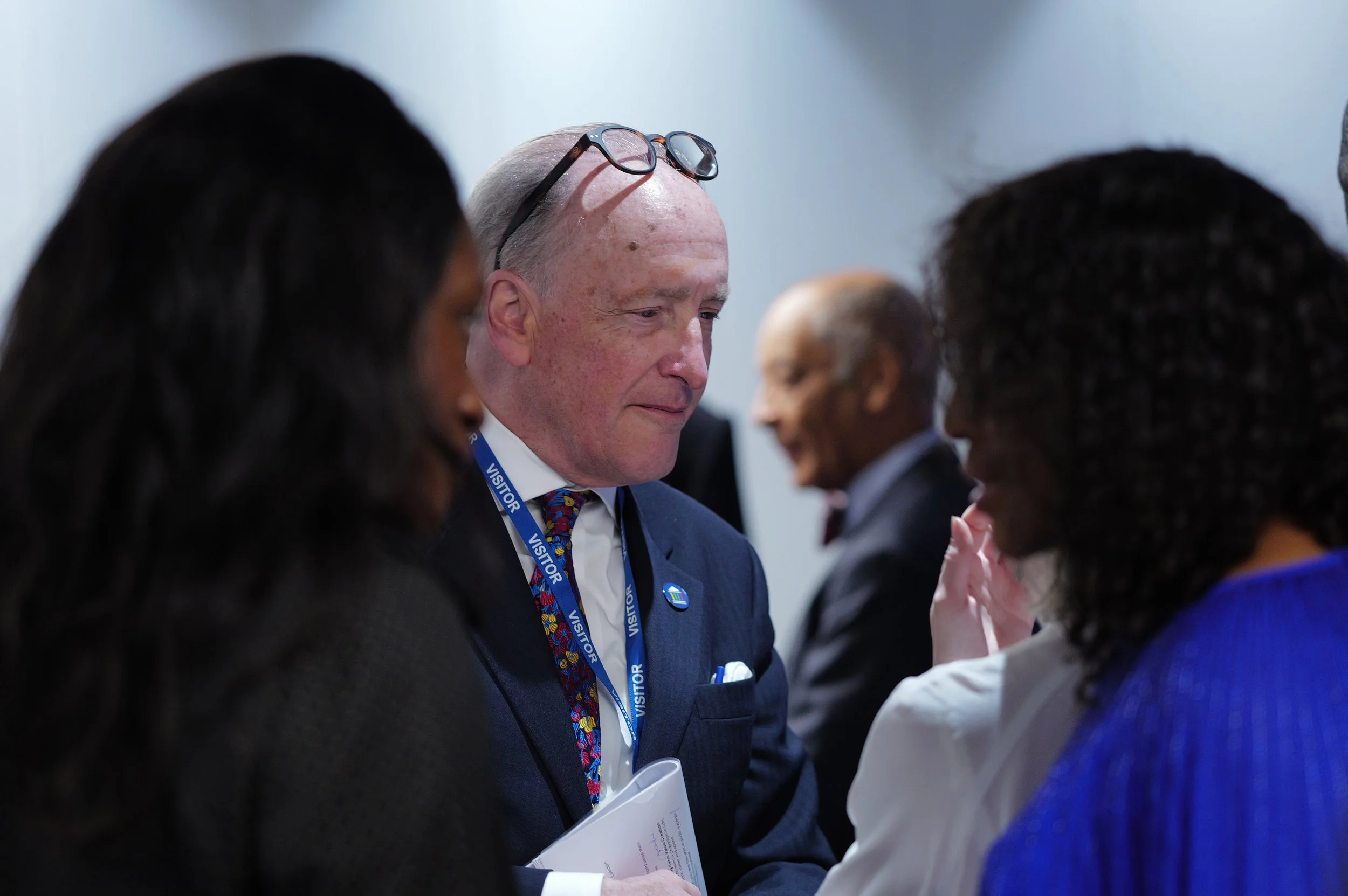Man in a suit with glasses on his head and a visitor badge talks with two women in a dimly lit setting.