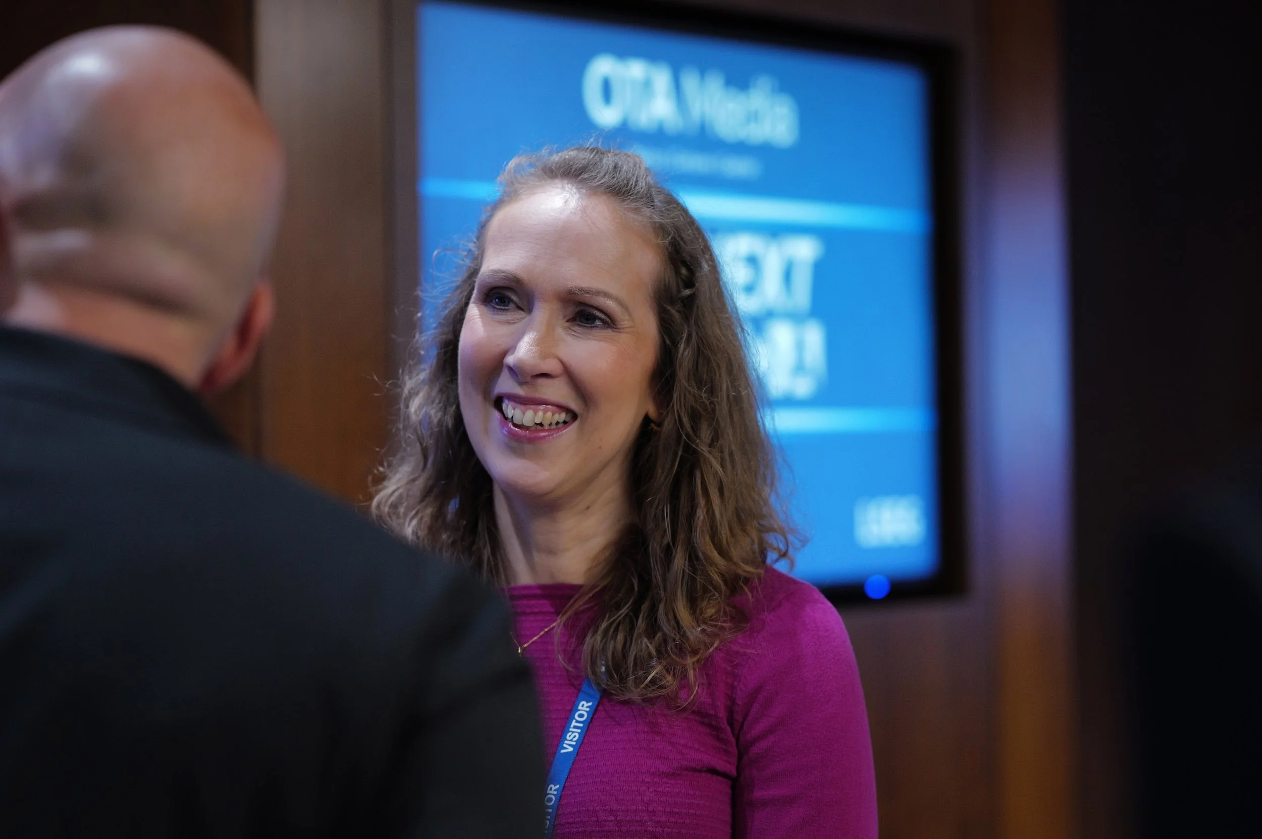 Smiling woman with curly hair wearing a magenta top and visitor badge talking to a bald man.