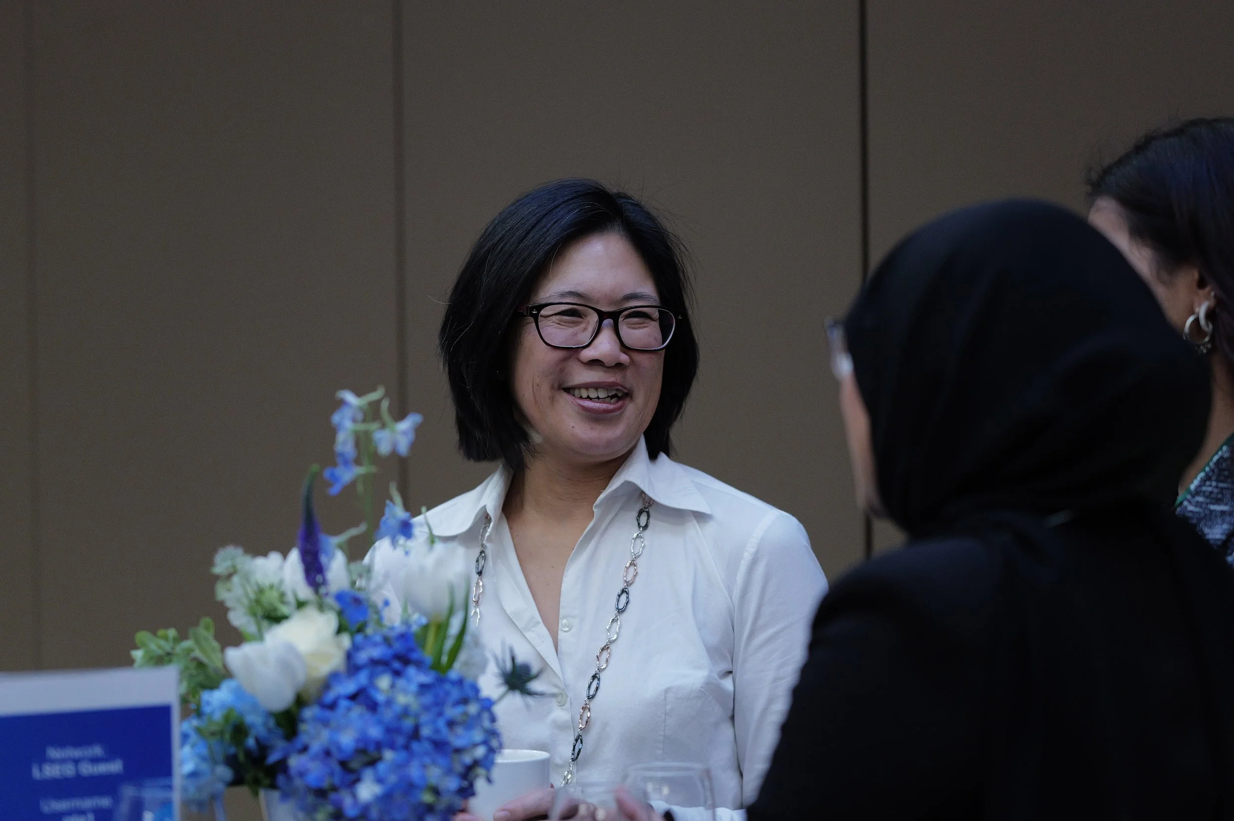 Smiling woman with short black hair and glasses wearing a white shirt converses with two people, one wearing a black hijab, near a table with blue and white flowers.