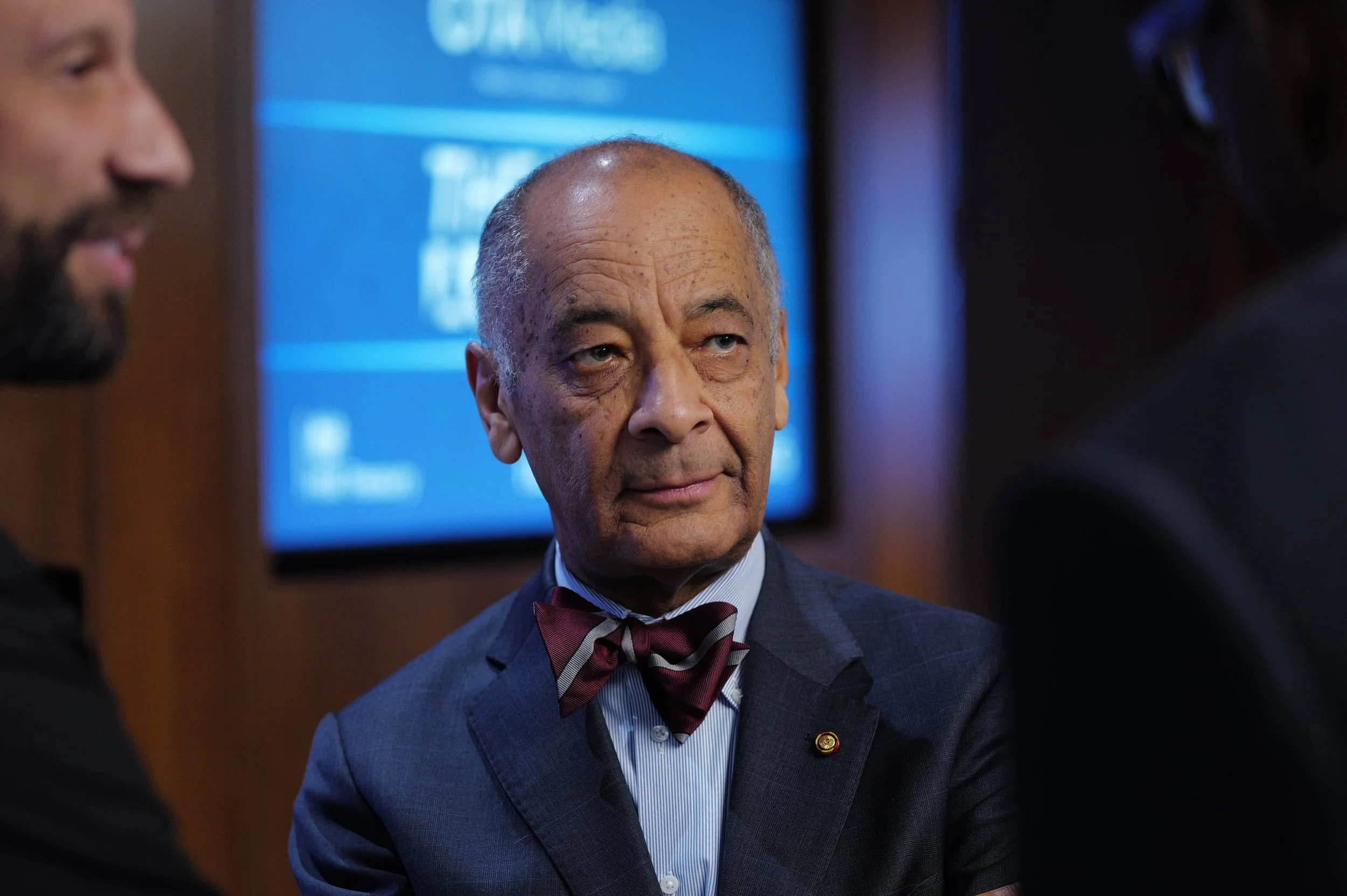 Older man in a blue suit with a striped shirt and red bow tie listening intently during a conversation at an indoor event.