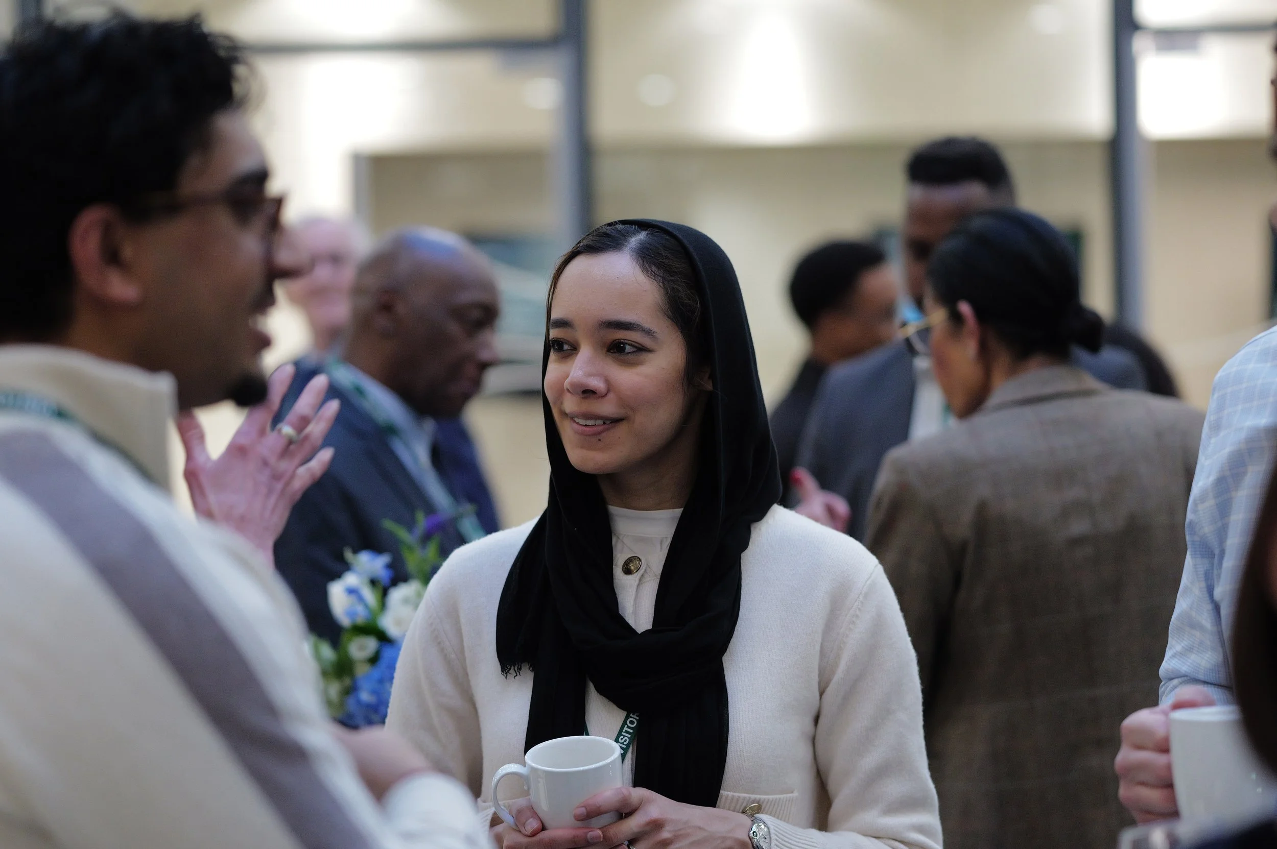 Young woman in a black headscarf and white sweater holding a white mug and smiling while talking to a man in glasses at a social event.