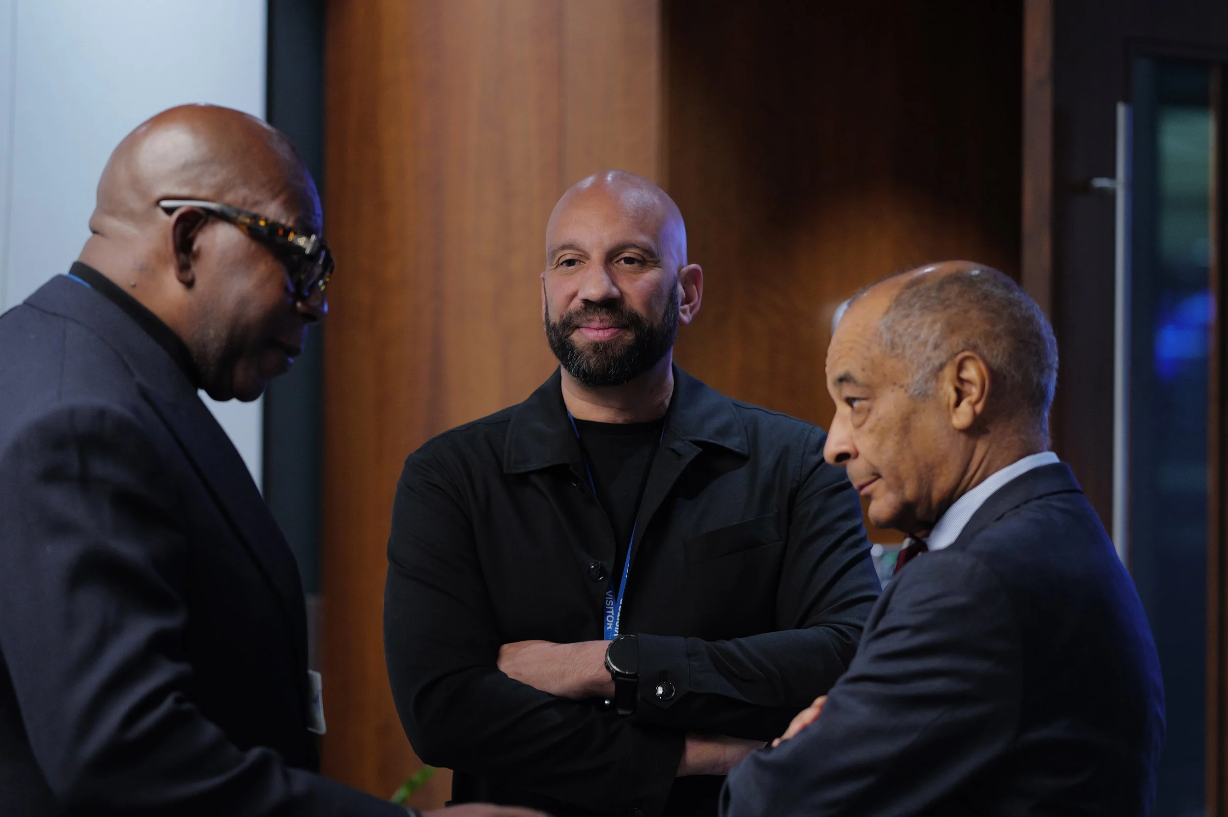 Three men in conversation indoors, one in the center with a beard and black jacket, the others in suits.