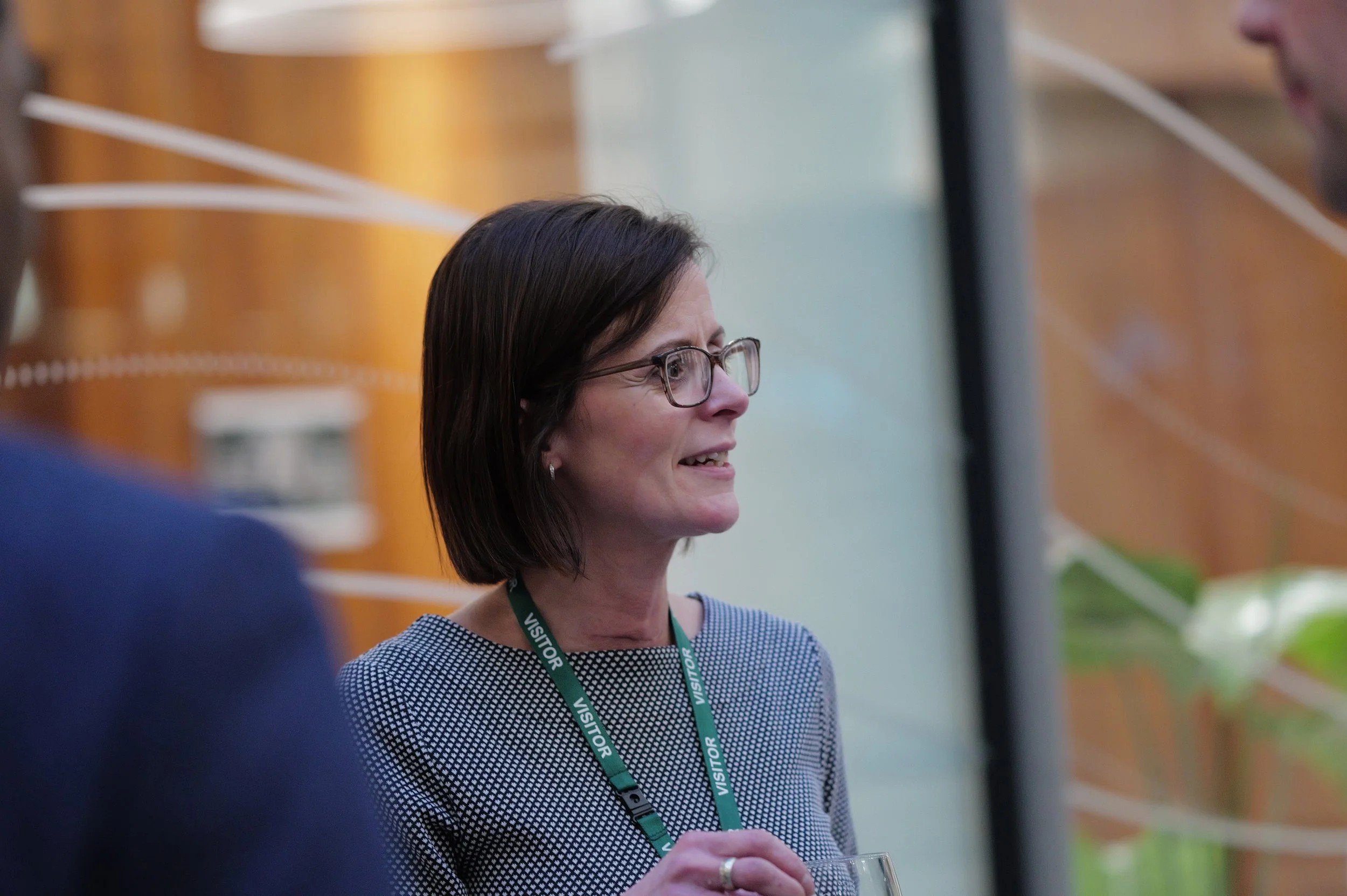 Woman with glasses wearing a visitor lanyard and holding a glass, engaged in conversation indoors.