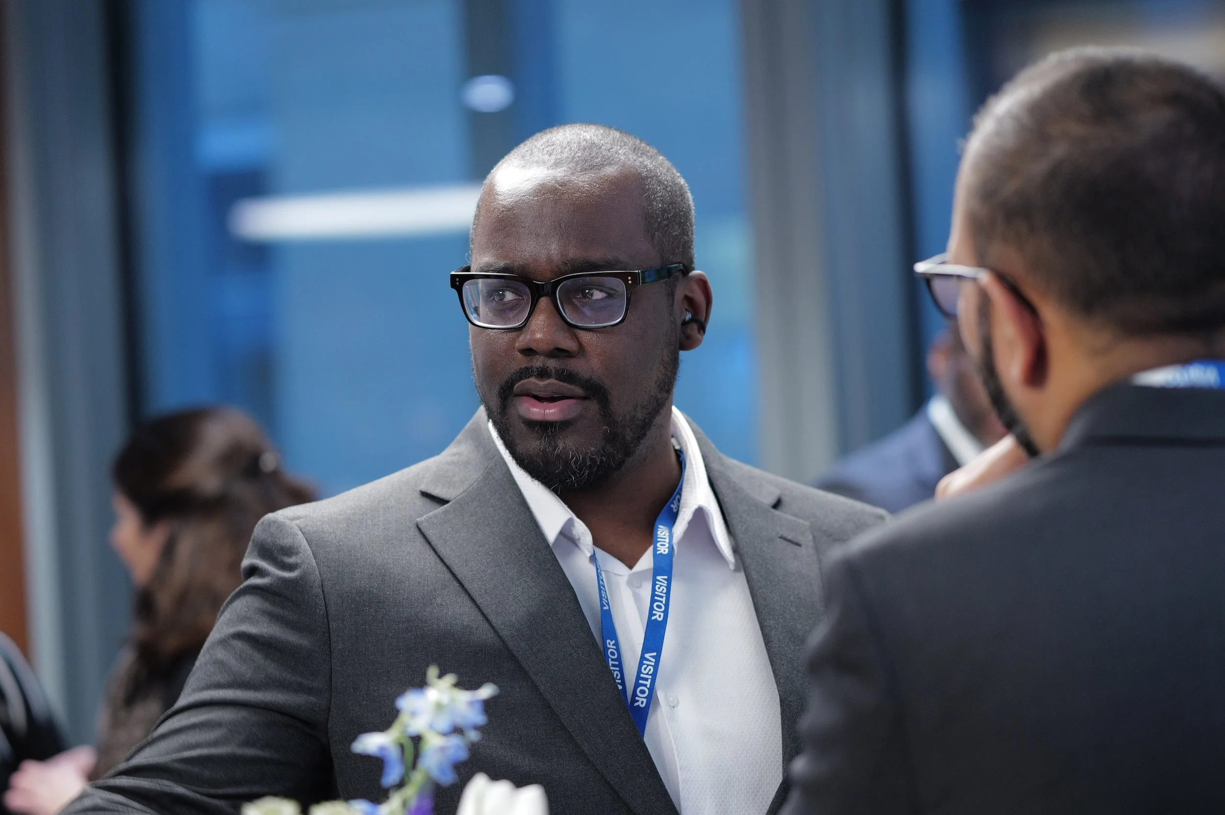 Man wearing glasses and a gray suit with a visitor badge, engaged in conversation at an indoor event.