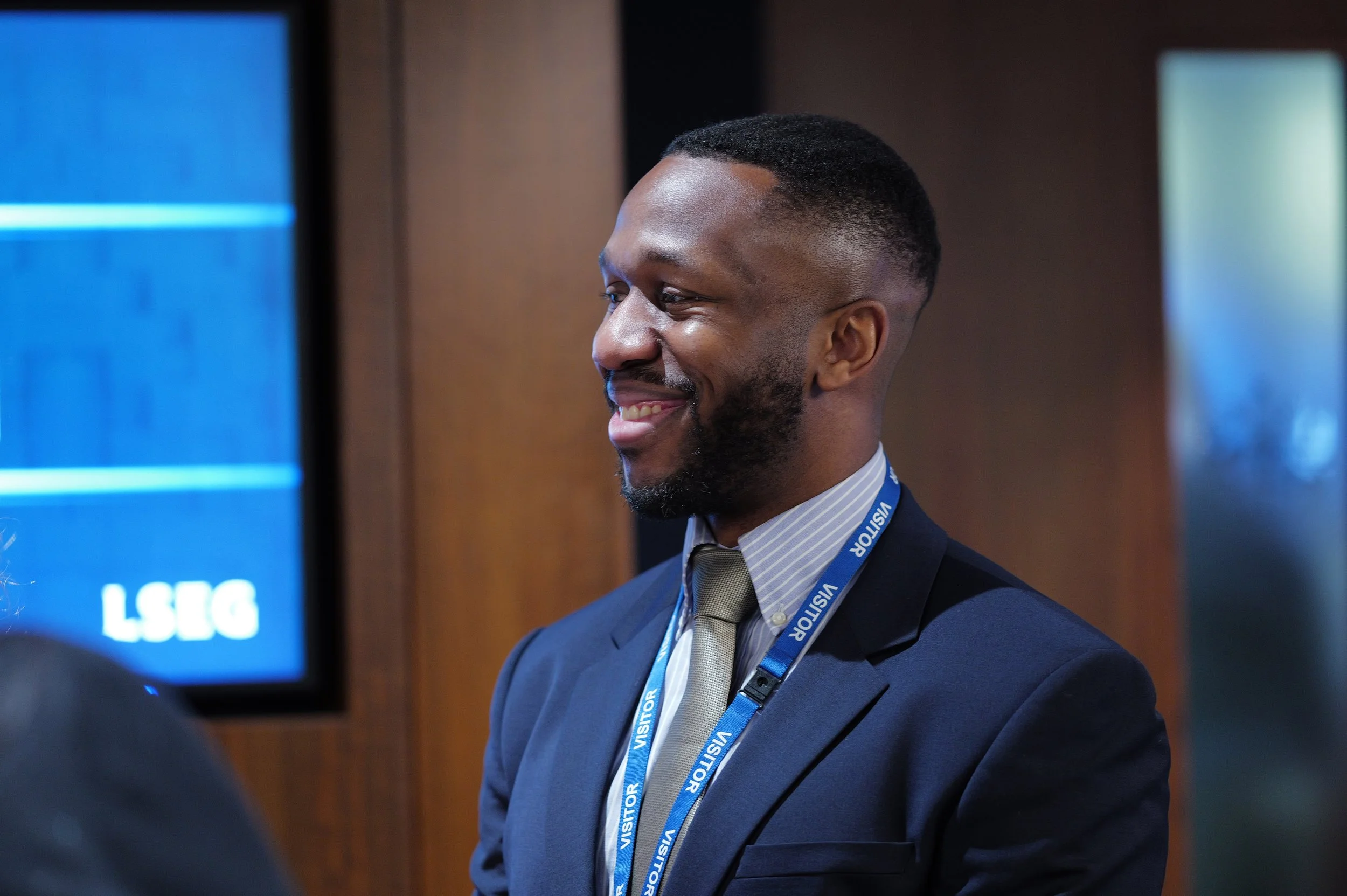 Smiling man in a suit with visitor badge standing indoors against a wooden wall.