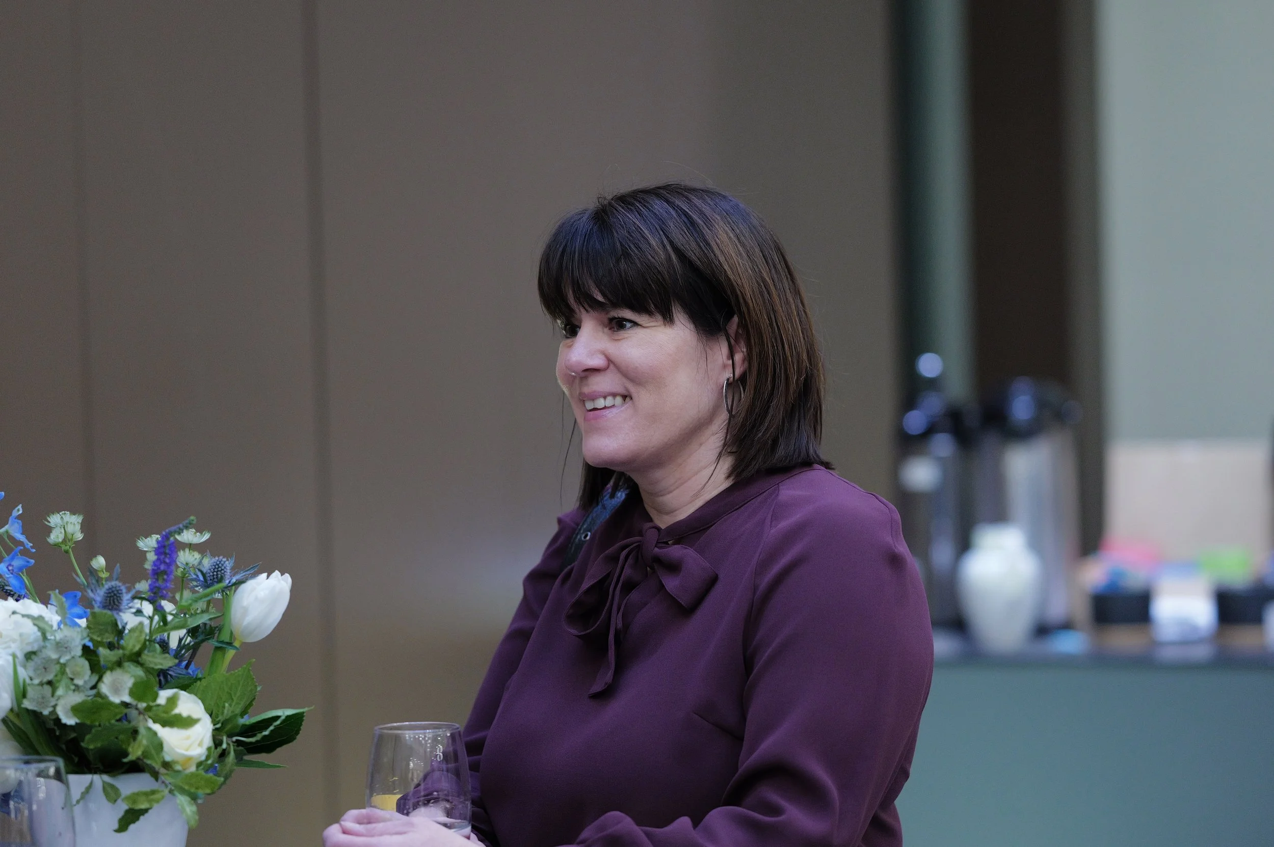Woman smiling and holding a glass, wearing a purple blouse with a bow at the neck, sitting near a vase of flowers.