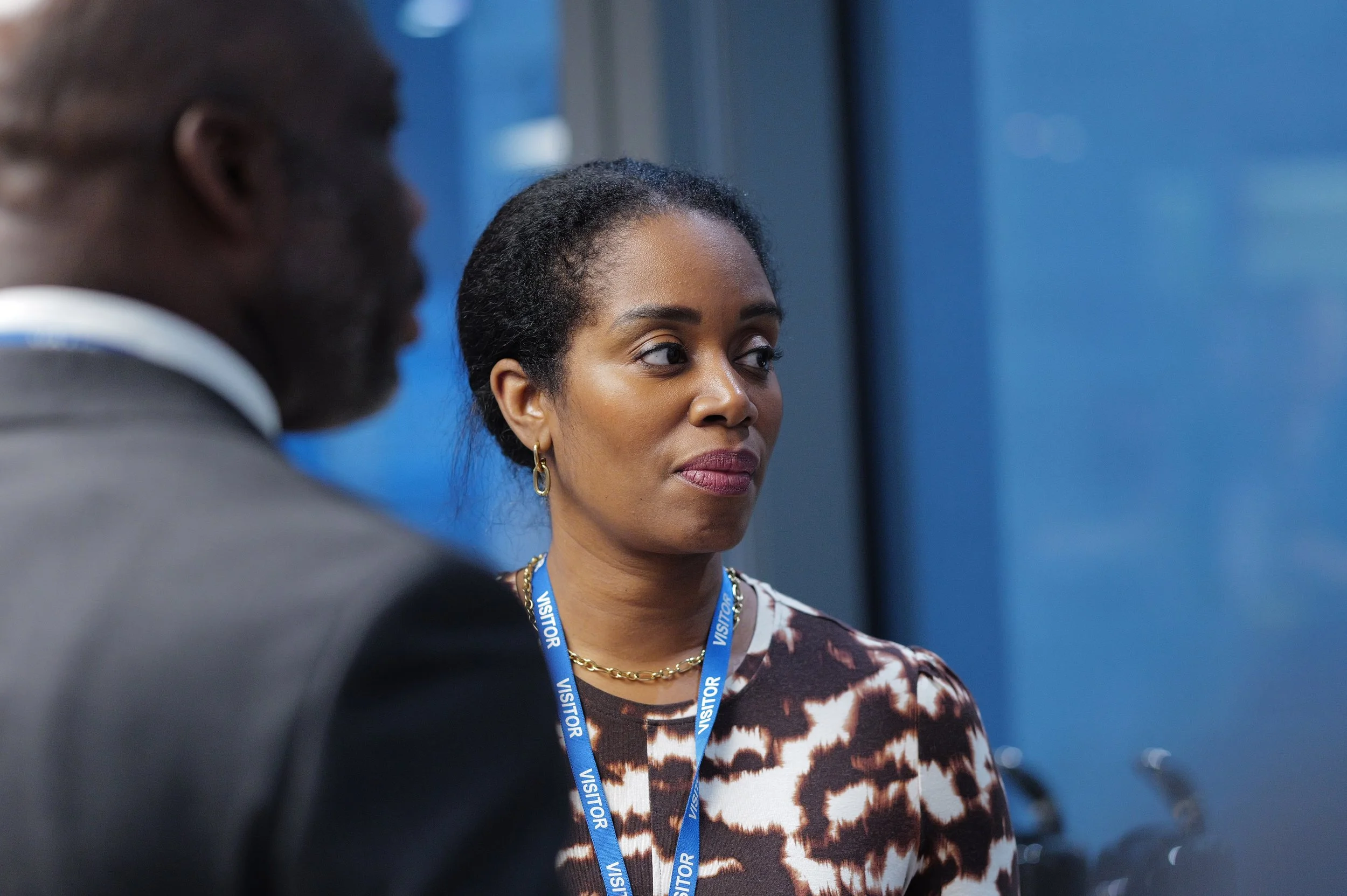 Woman wearing a visitor badge and patterned shirt looking to the side, with a man in a suit blurred in the foreground.