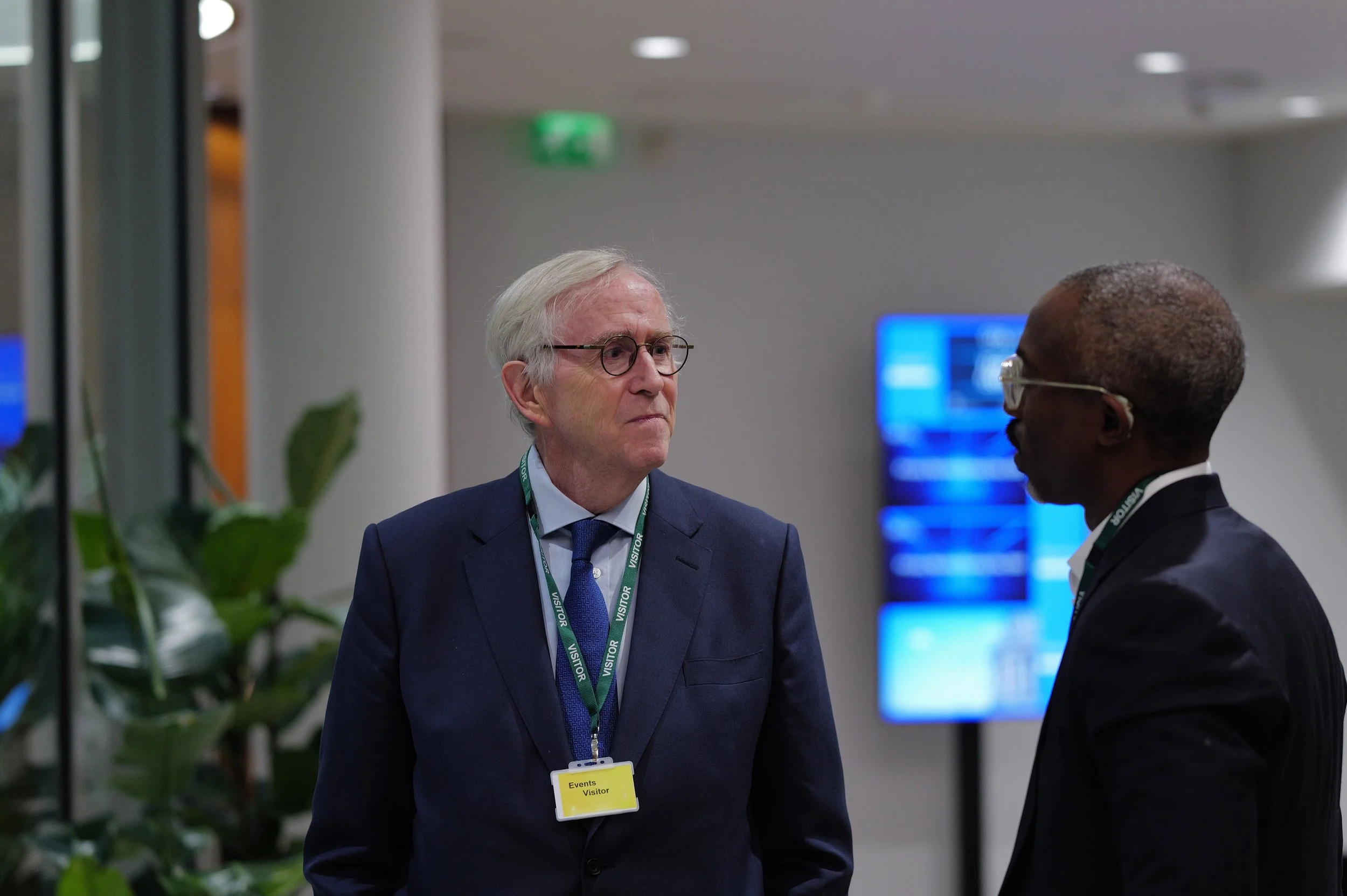 Two men in suits having a conversation indoors, with one wearing glasses and a visitor badge.