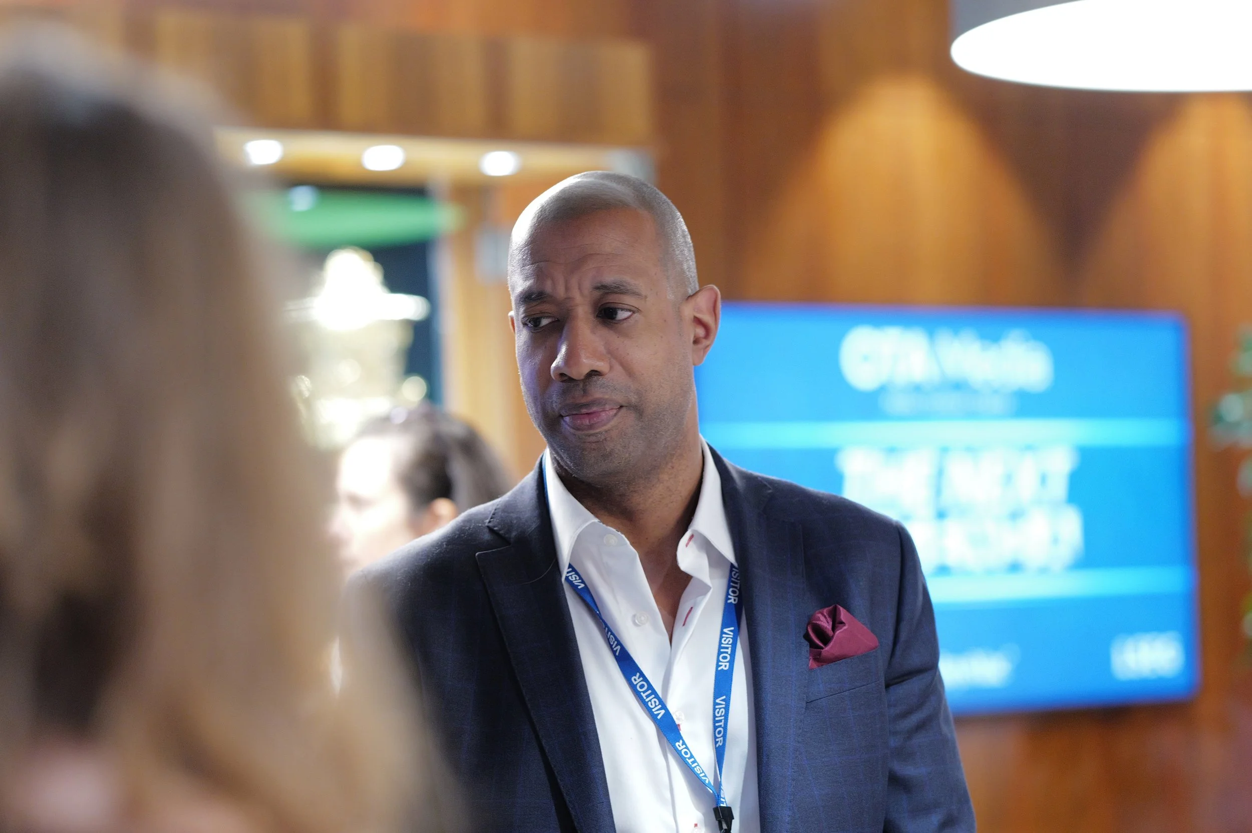 Man in a suit with a visitor badge looking to the side in a professional setting with a blurred person in the foreground.
