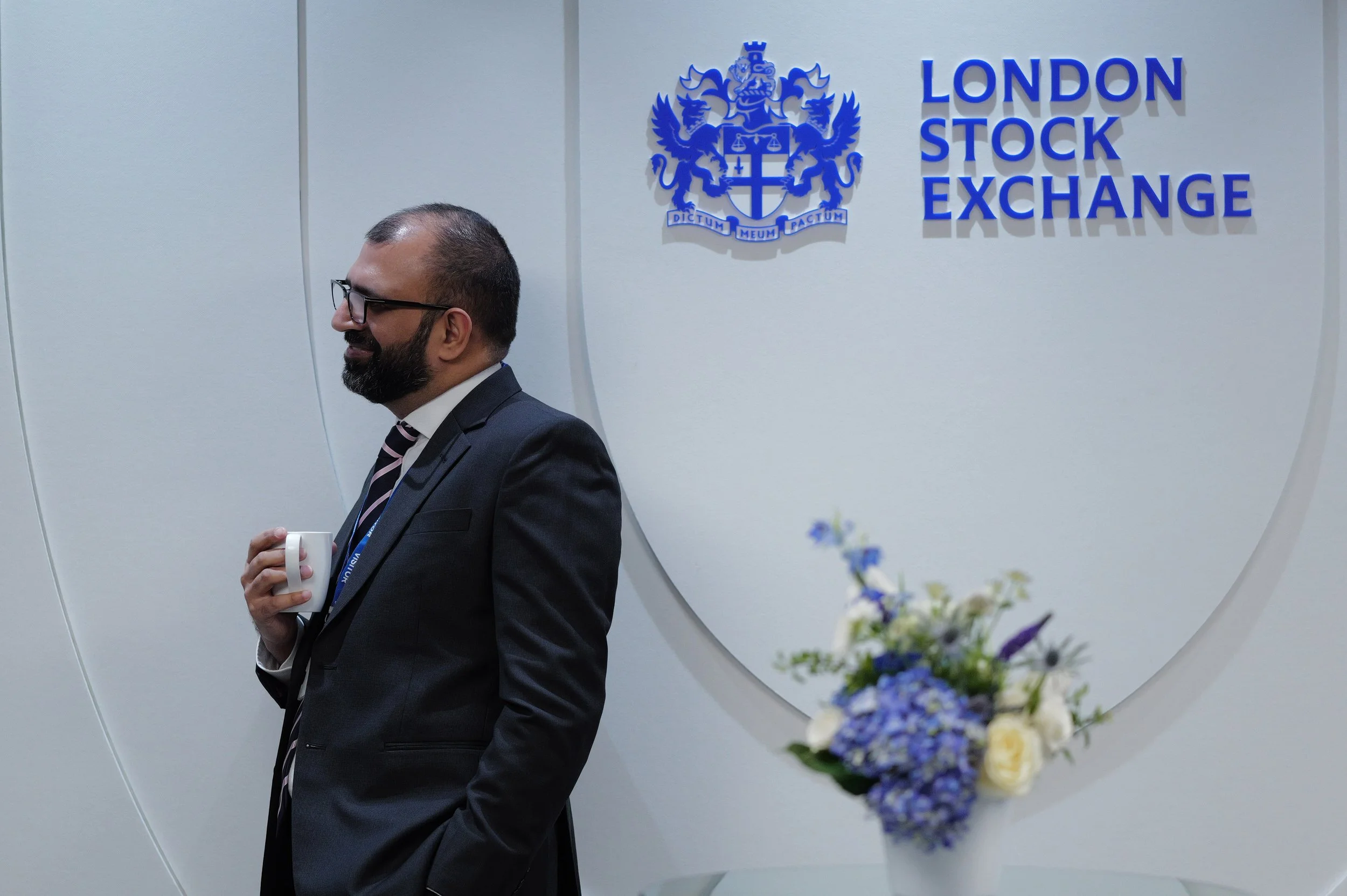 Man in glasses and suit holding a mug, standing near a wall with the London Stock Exchange logo.