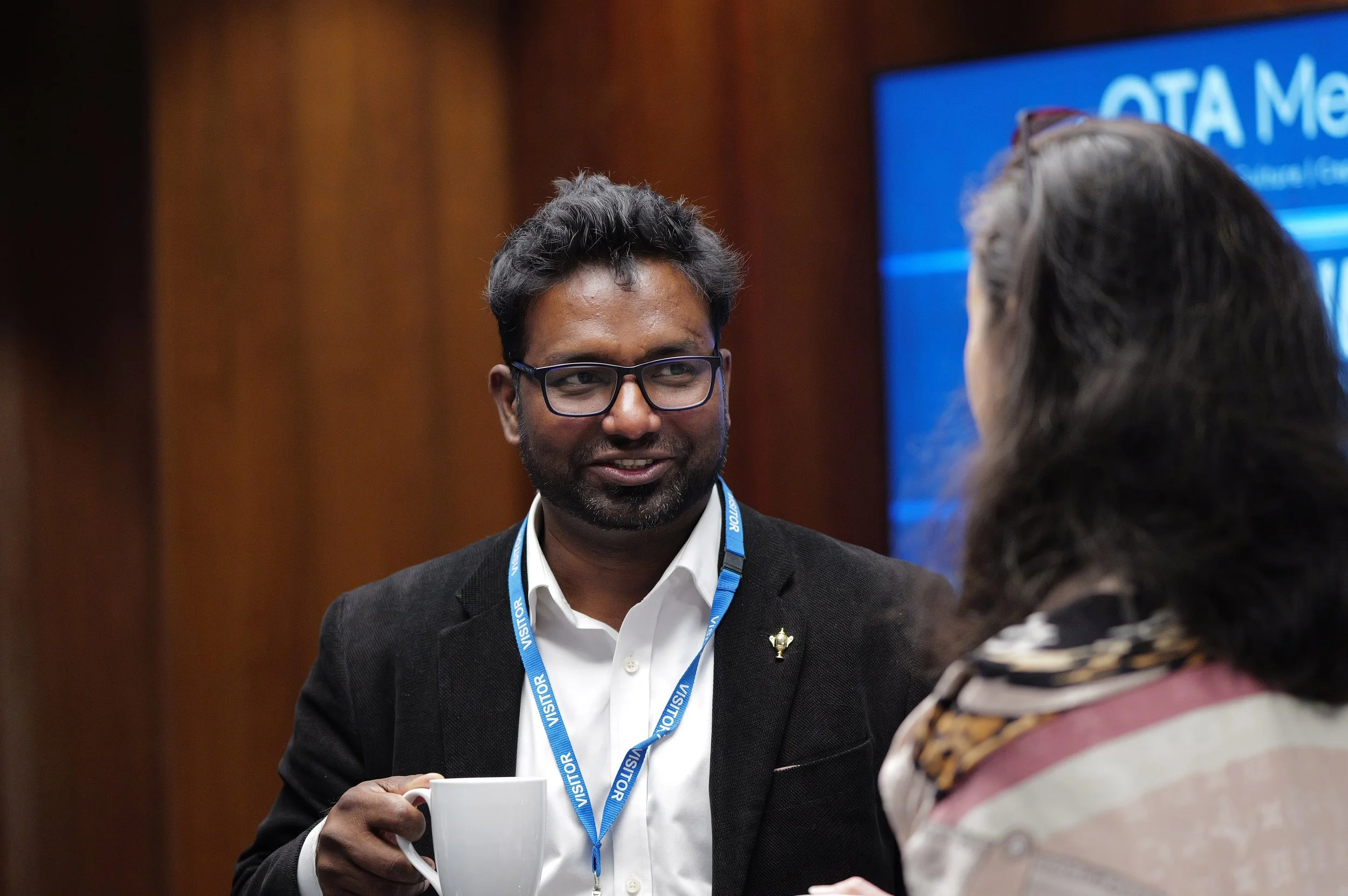 Man wearing glasses and a visitor badge holding a white cup, talking to a woman with long hair and a patterned scarf.