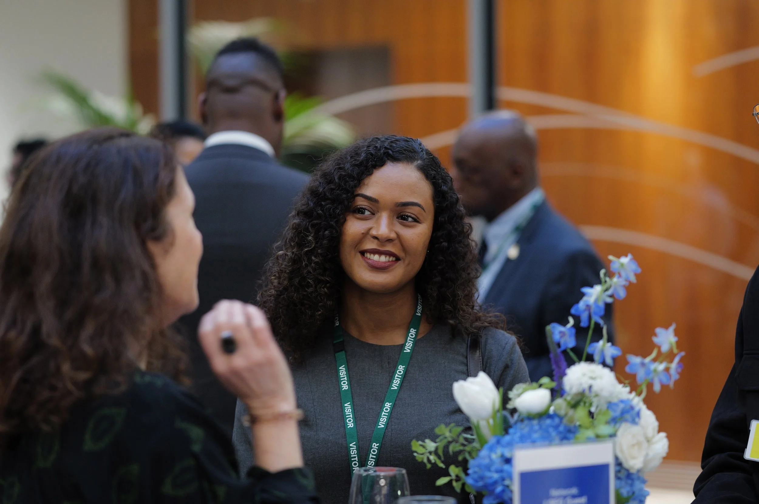 Smiling woman with curly hair wearing a visitor badge, engaged in conversation at a professional event.